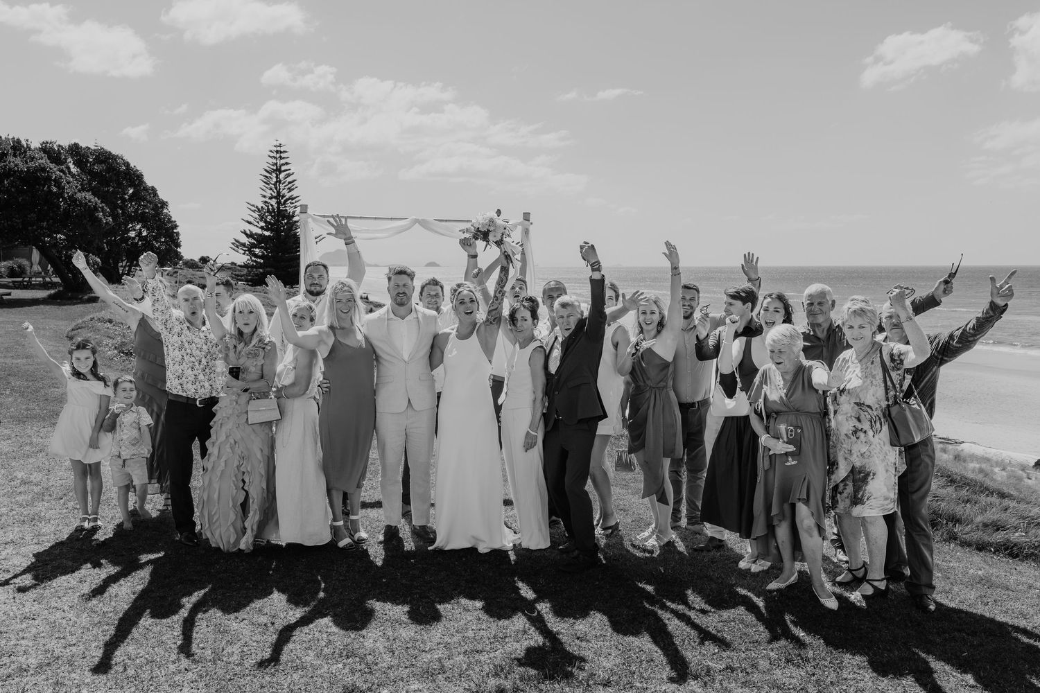 Black and white wedding photo of a large group celebrating outdoors near the ocean, with hands raised in joy.
