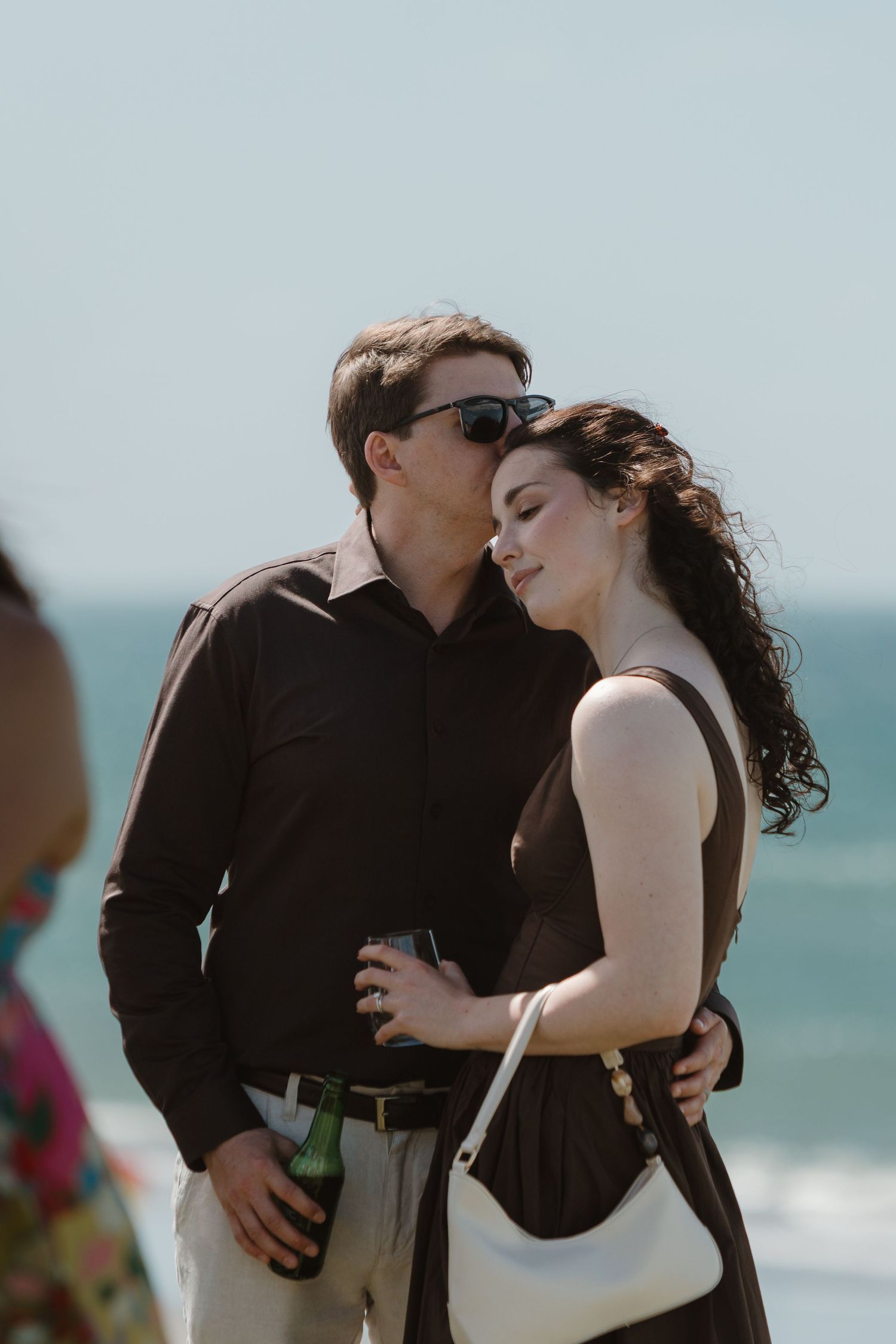 A romantic couple embraces on a sunny beach, man in black shirt holding a beer bottle, woman in brown dress with white bag.