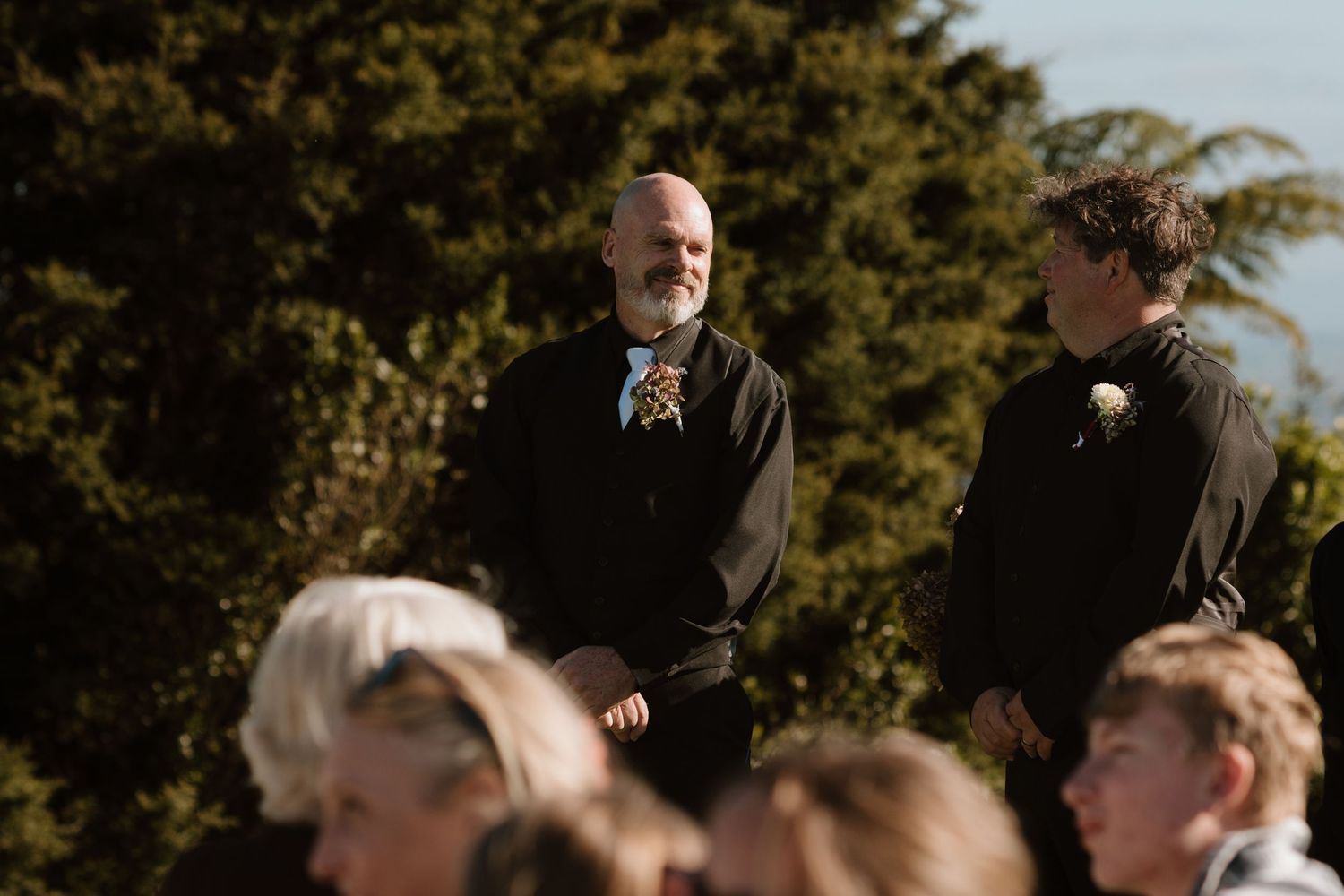 People gathered outdoors at a formal event on a sunny day with trees visible in the background.