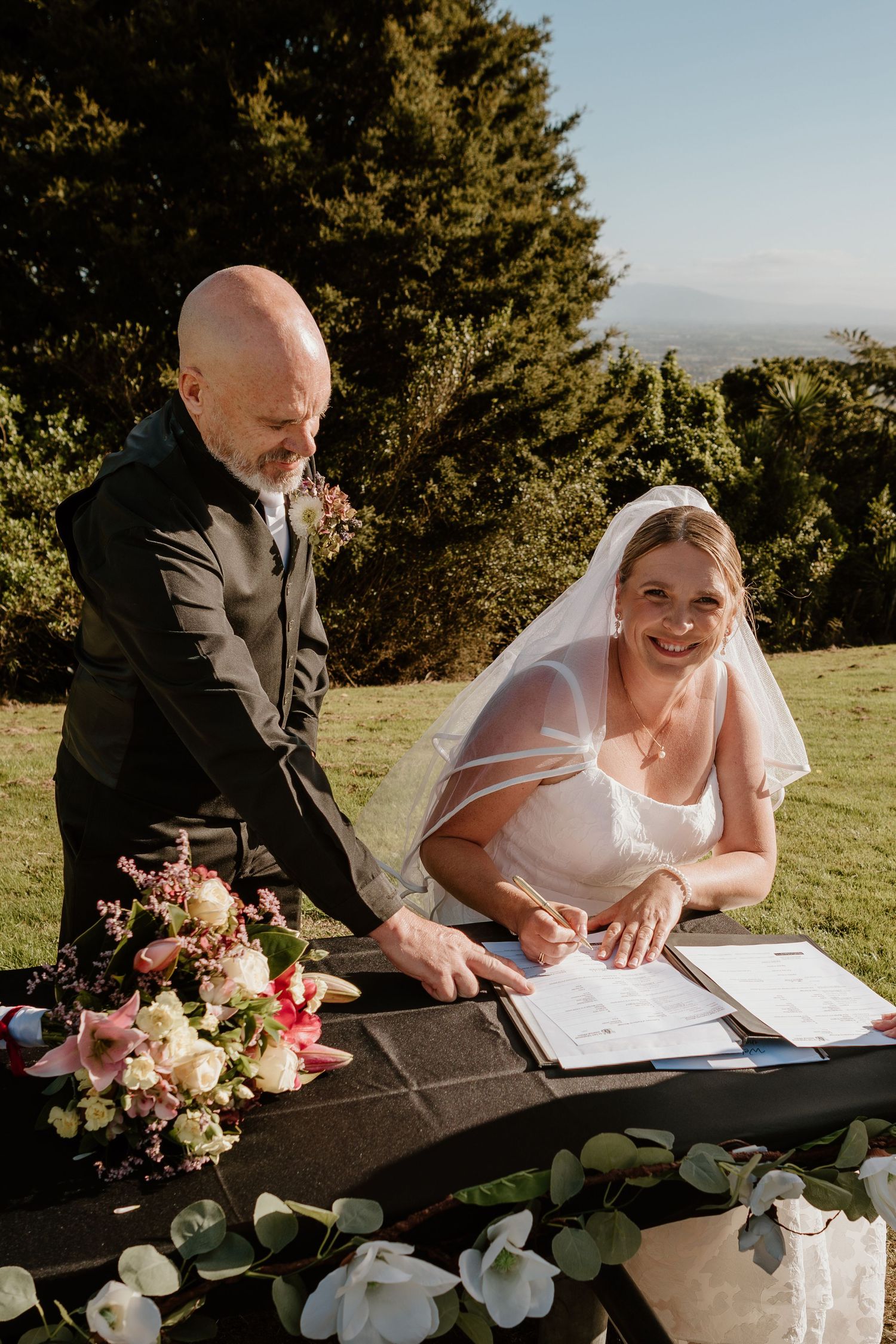 A couple signing their wedding documents at an outdoor ceremony with floral arrangements and greenery.