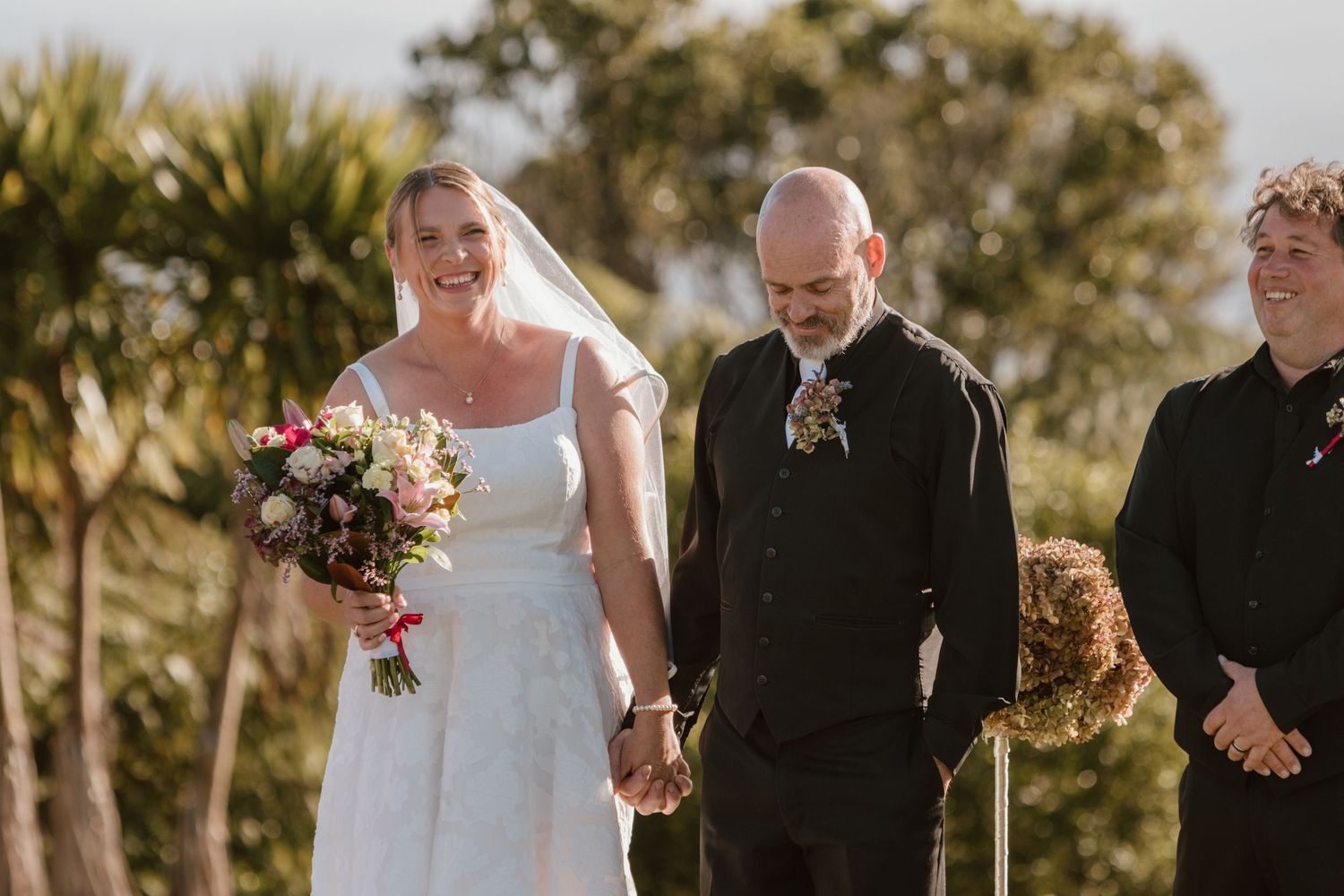 A bride and groom exchange vows during an outdoor wedding ceremony with guests standing nearby at golden hour.