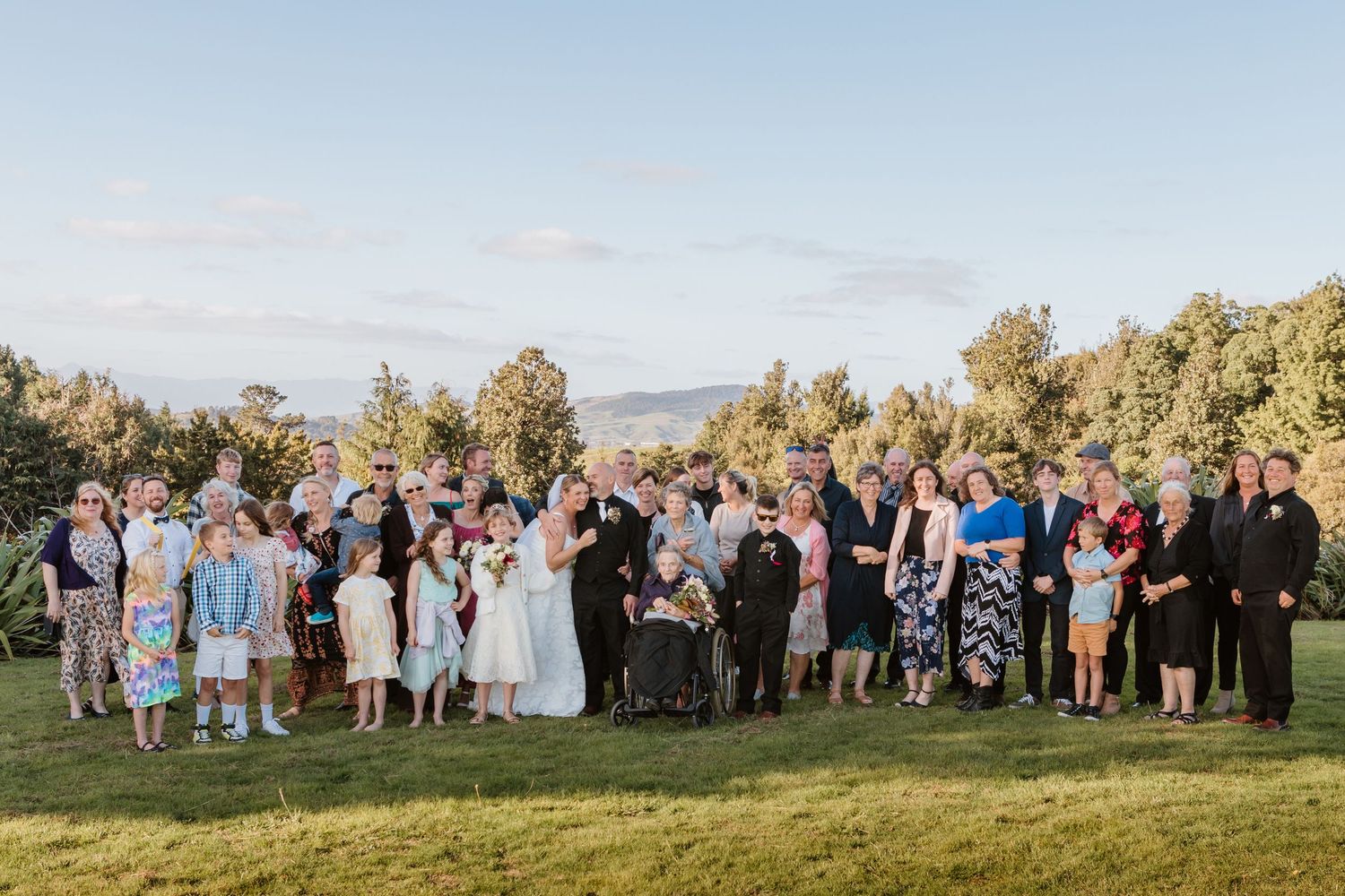 Wedding guests gather on a green lawn with mountains and trees in the background at an outdoor celebration.