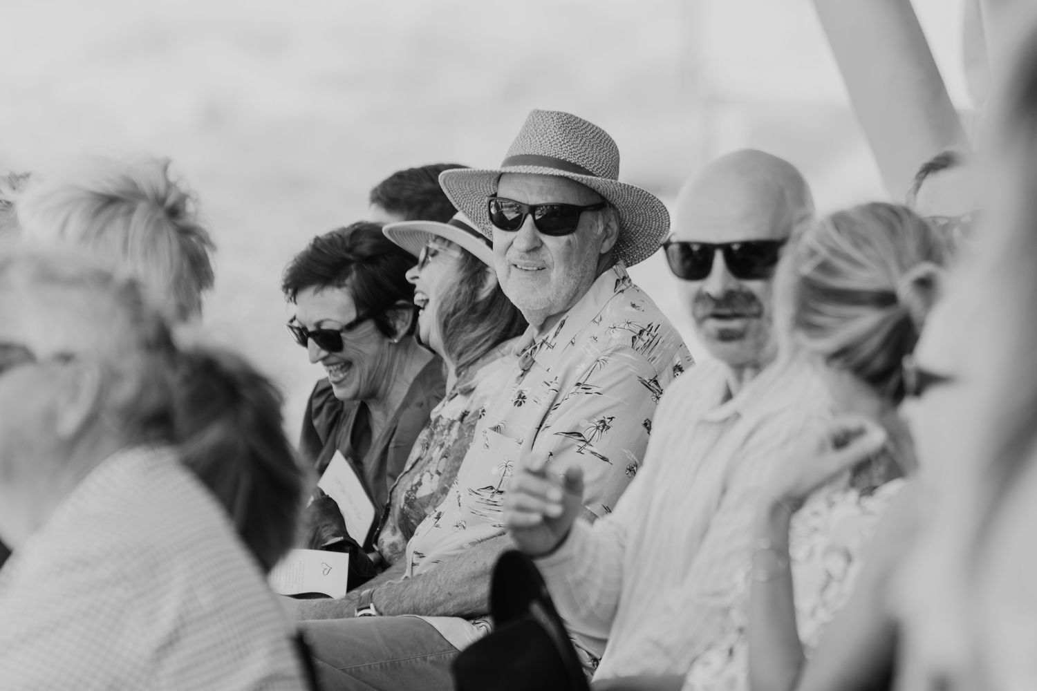Wedding guests in sunhats and summer attire seated outdoors in black and white photography. Papamoa Surf Lifesaving Club Wedding