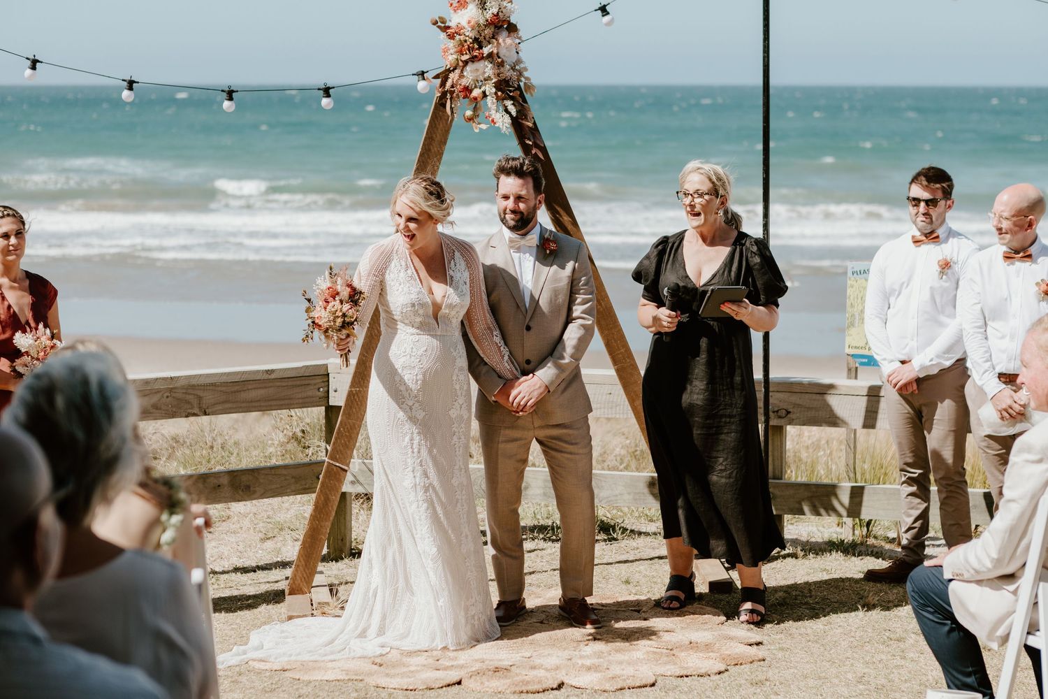 Beachside wedding ceremony setup with wooden arch and string lights overlooking the ocean. Papamoa Surf Lifesaving Club Wedding