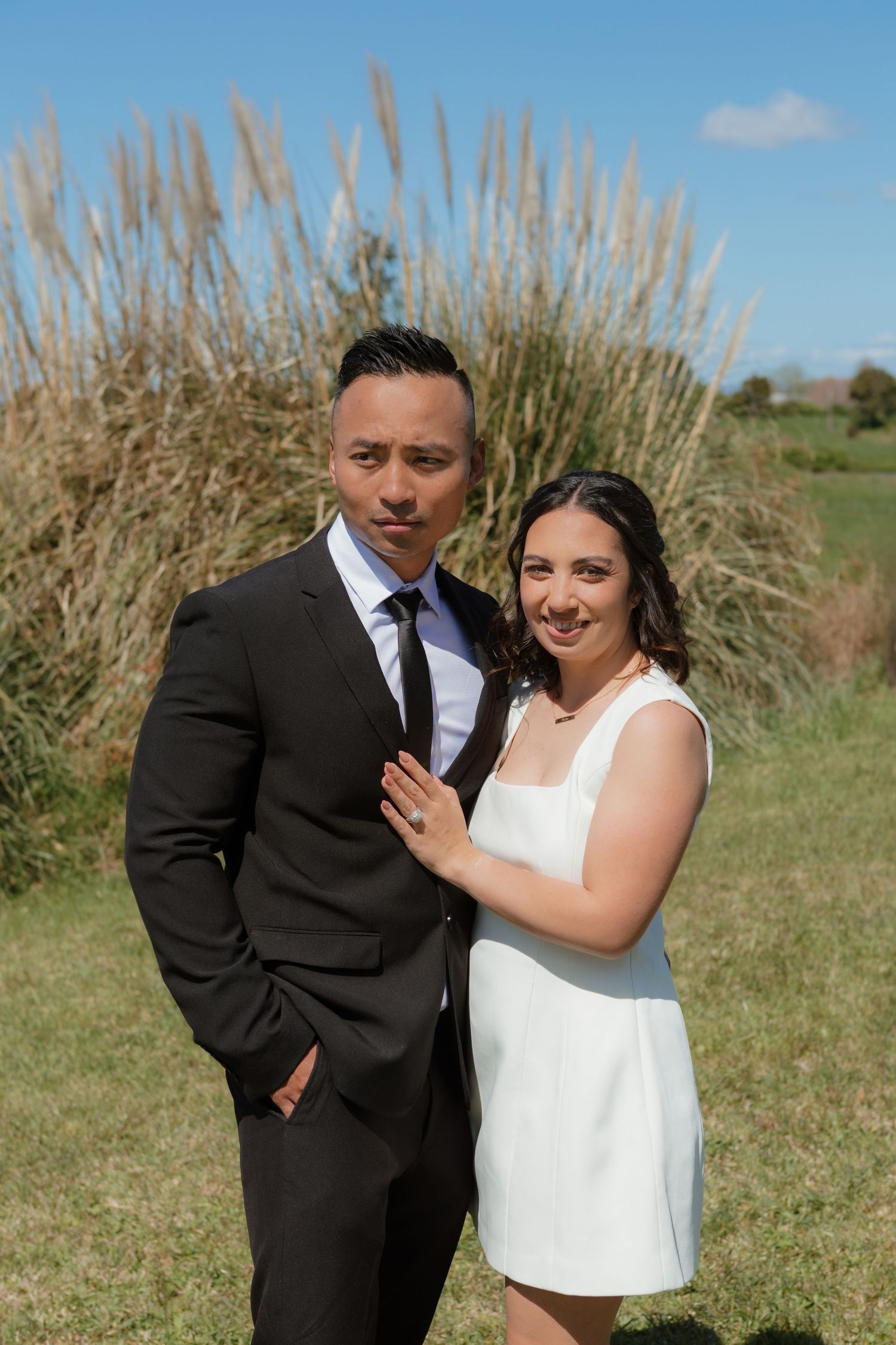 A couple in formal attire poses together outdoors against tall ornamental grass on a sunny day.