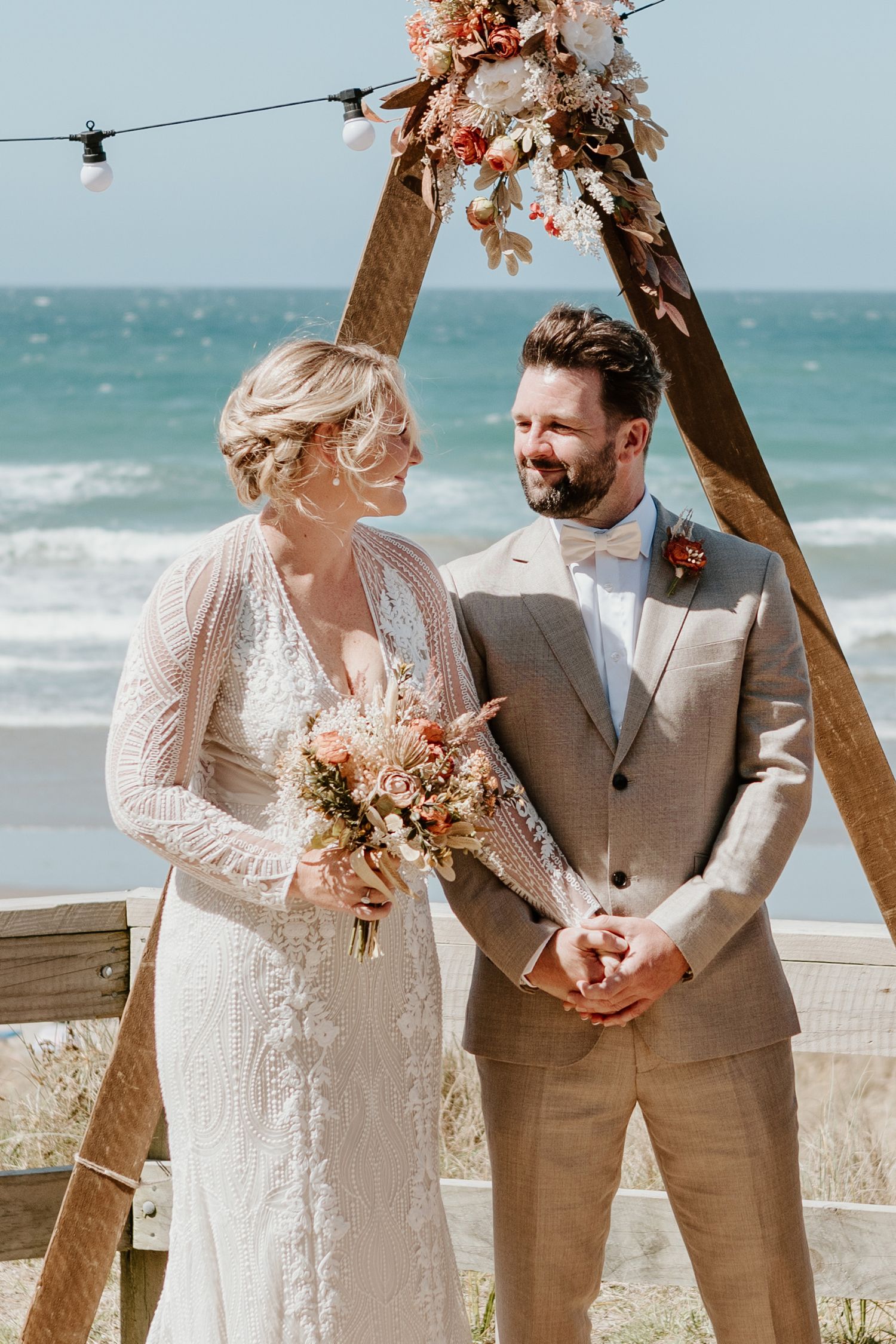 Beach wedding ceremony under wooden arch with dried floral decorations and string lights overlooking turquoise ocean.