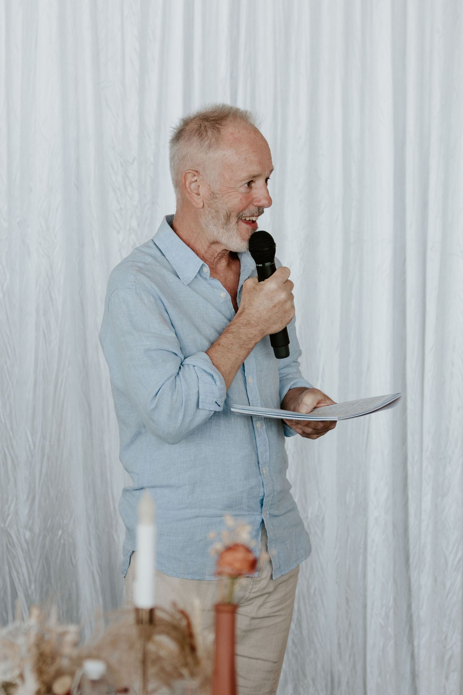 A cheerful person in a light blue button-down shirt stands with a microphone and paper at an event against a white curtain.
