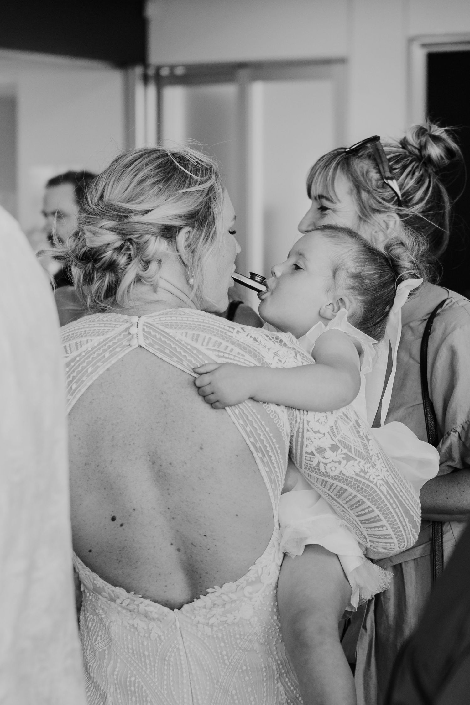 A black and white candid moment shows a toddler in white embracing someone in a backless lace dress.