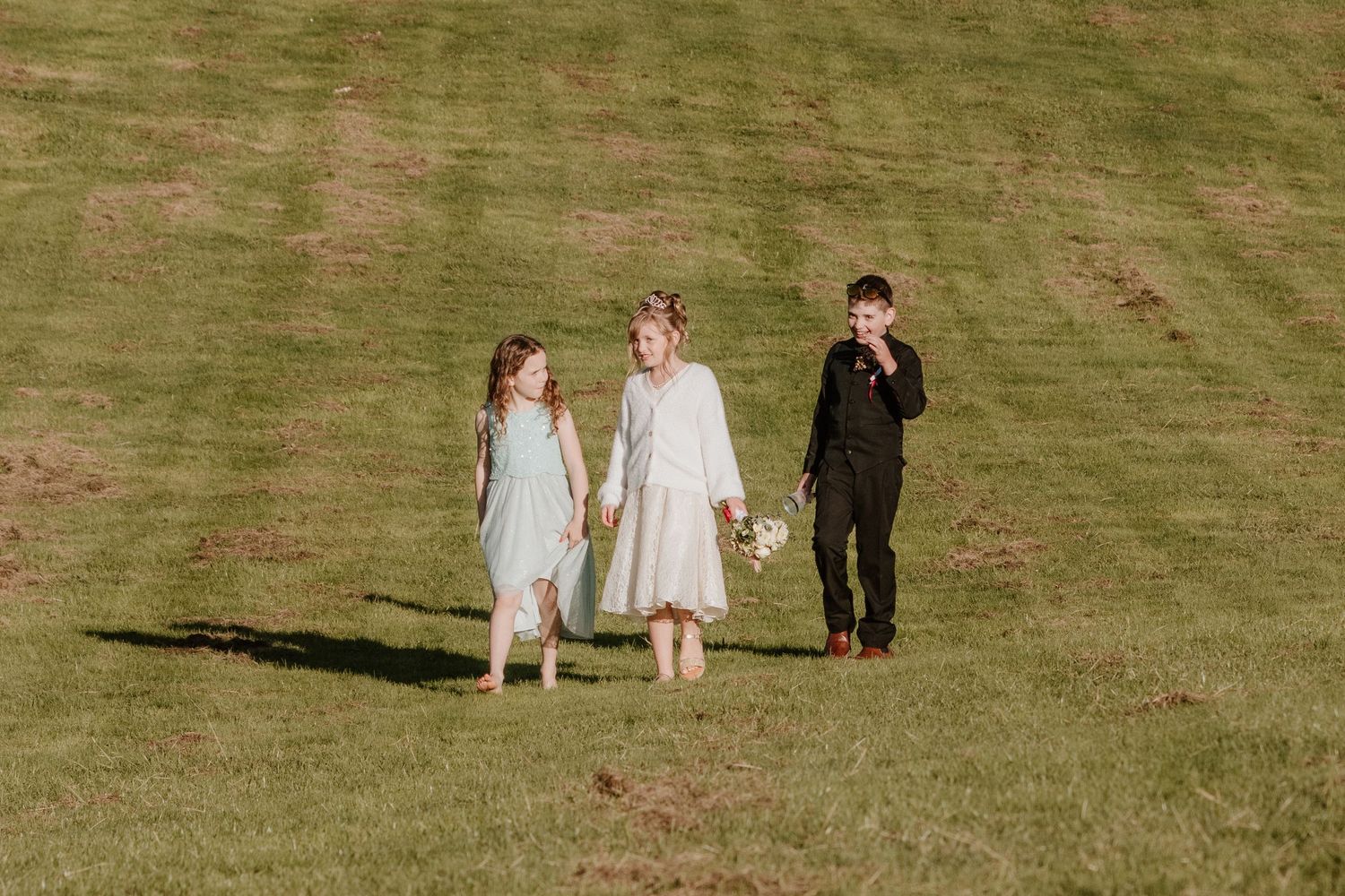 Three figures in formal attire walk together across a sunlit grassy hillside.