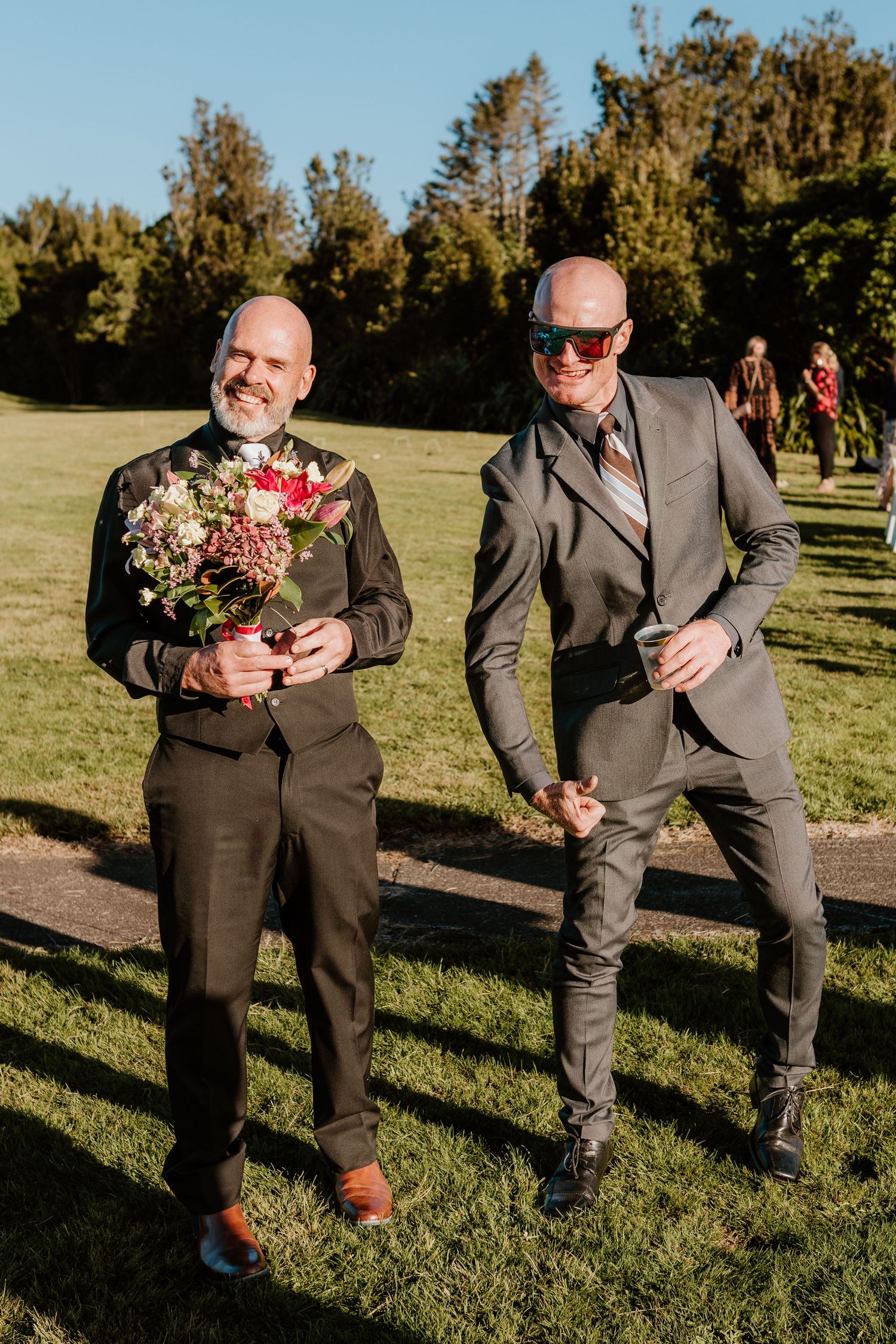 Two groomsmen in gray suits strike playful poses while holding wedding bouquets outdoors on a sunny day.