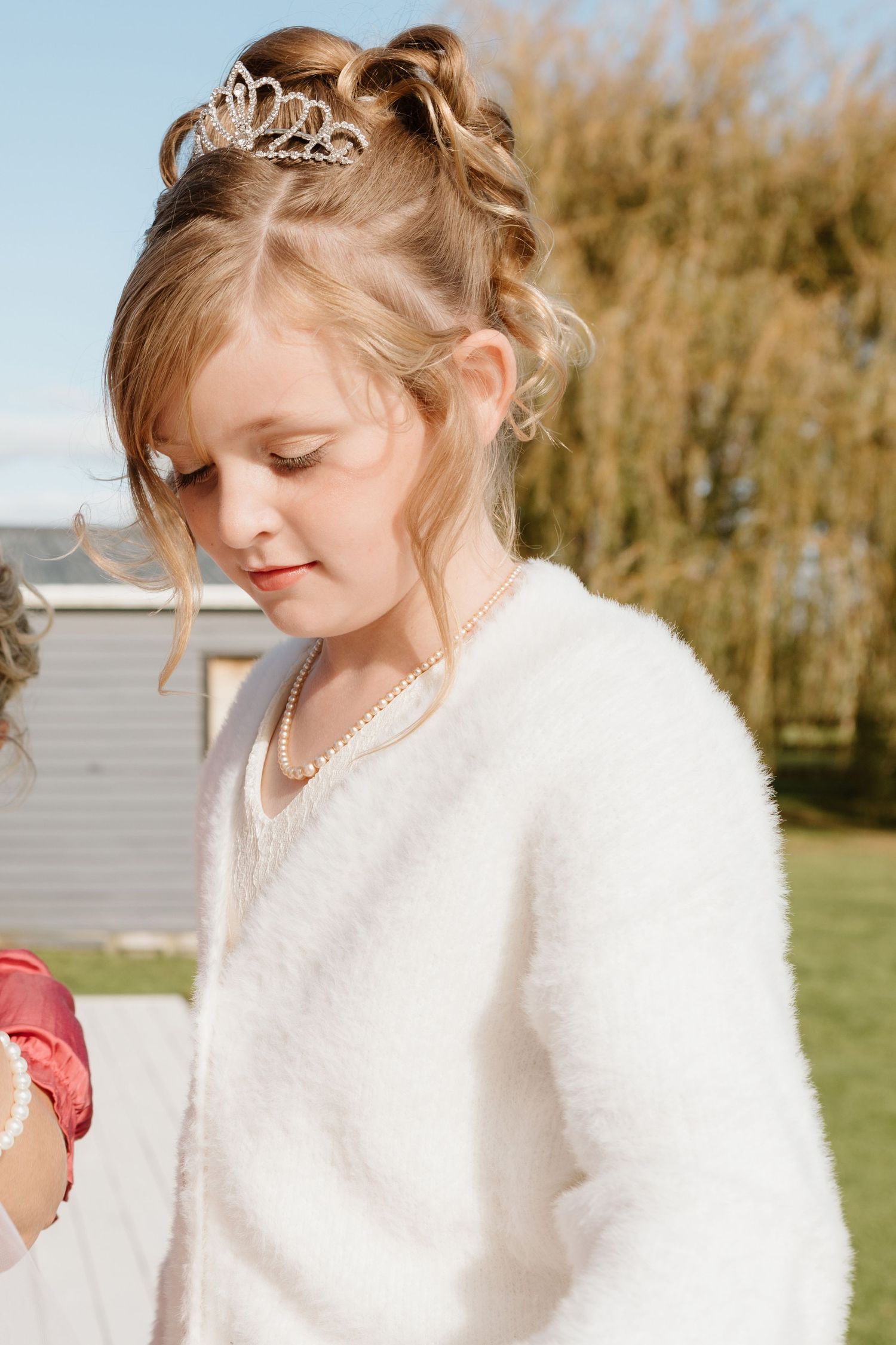A person in a white fuzzy sweater and delicate tiara wearing pearls stands in a garden setting on a sunny day.