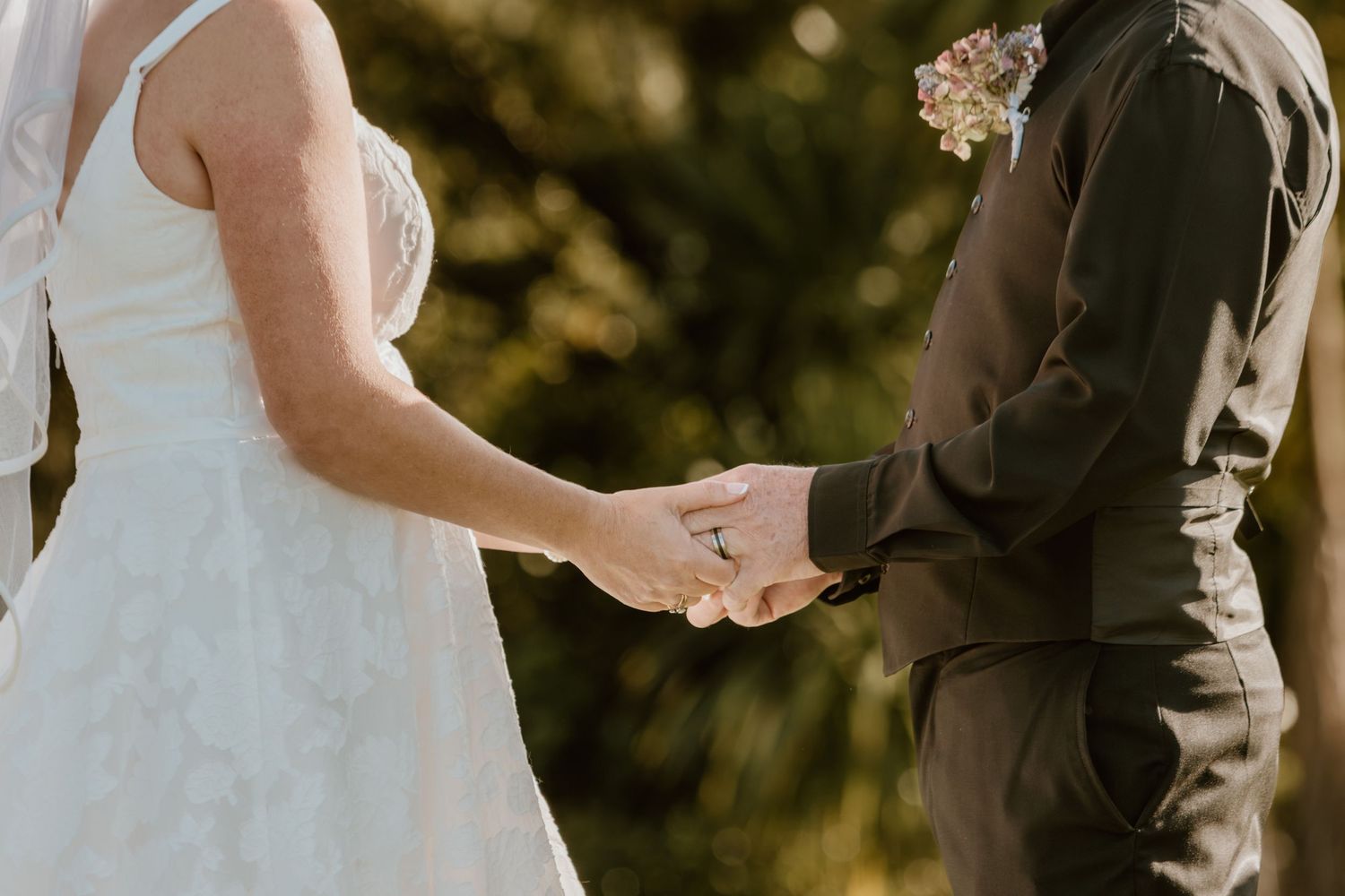 A tender moment at a wedding ceremony as a couple holds hands while wearing formal attire in golden sunlight.