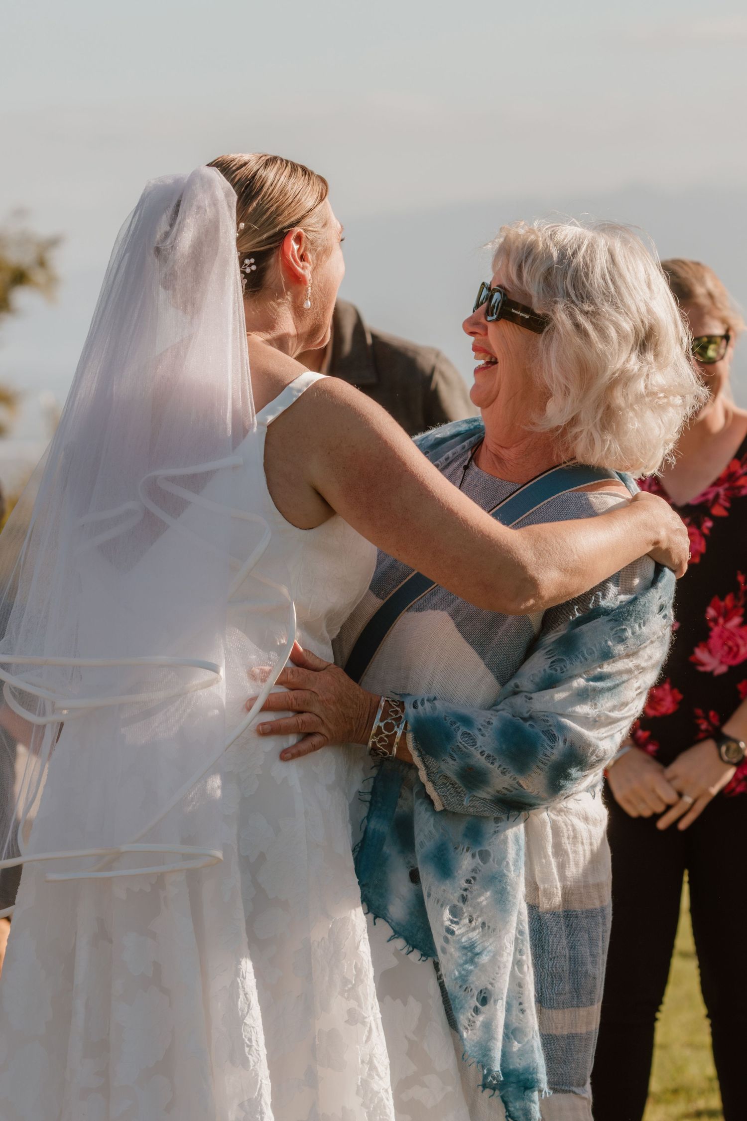A candid moment of a bride in a white wedding dress and veil embracing a guest in blue at an outdoor ceremony.