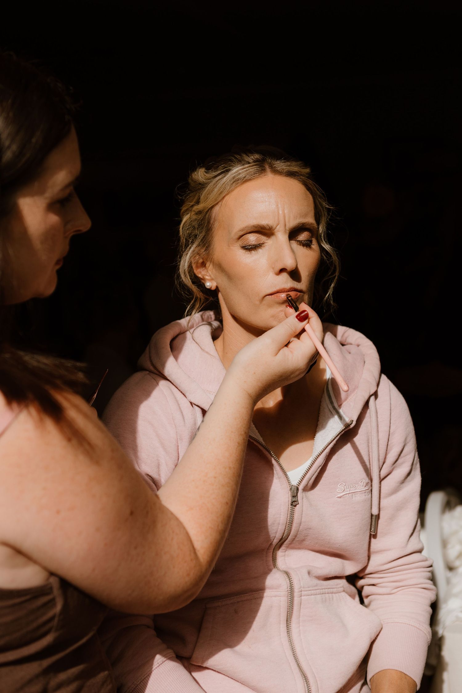 A makeup artist applies lipstick to someone wearing a pink zip-up hoodie in dramatic lighting.