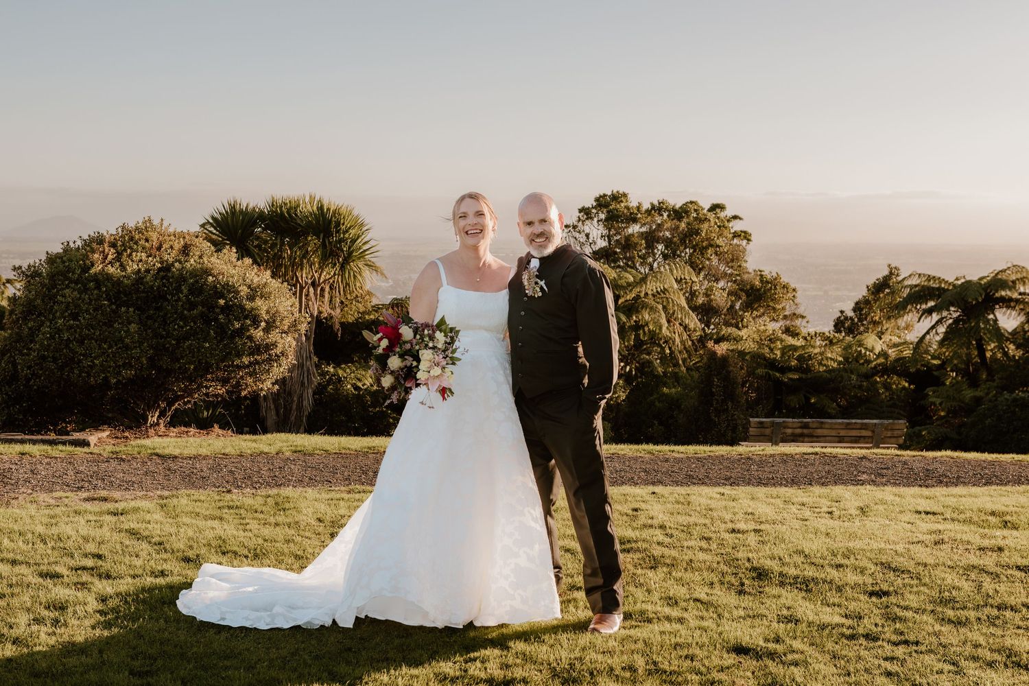 A couple in formal wedding attire poses together on a grassy hilltop during golden hour with trees in the background.