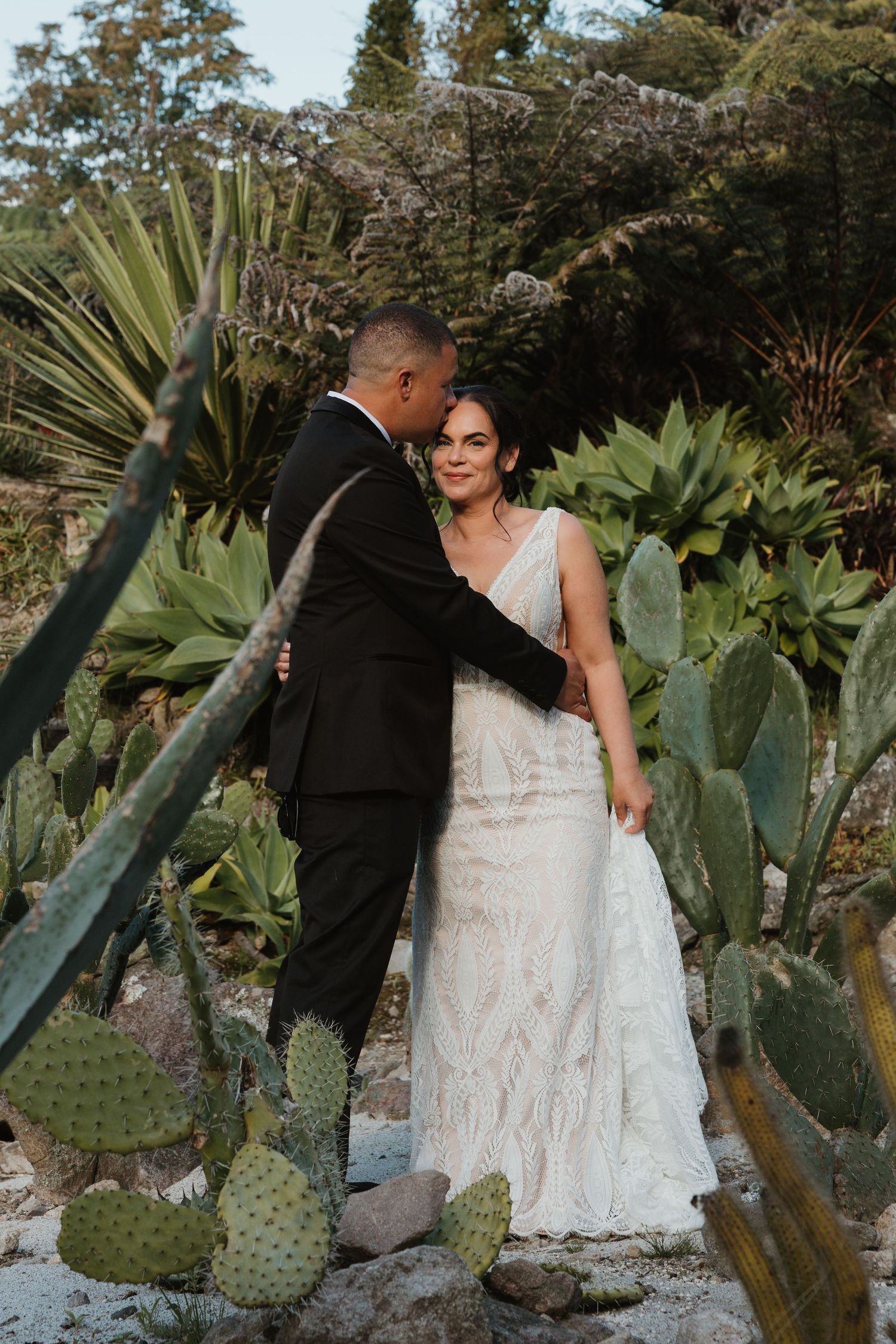 A couple shares romantic moments in a lush garden setting with cacti and tropical plants during their wedding photoshoot.