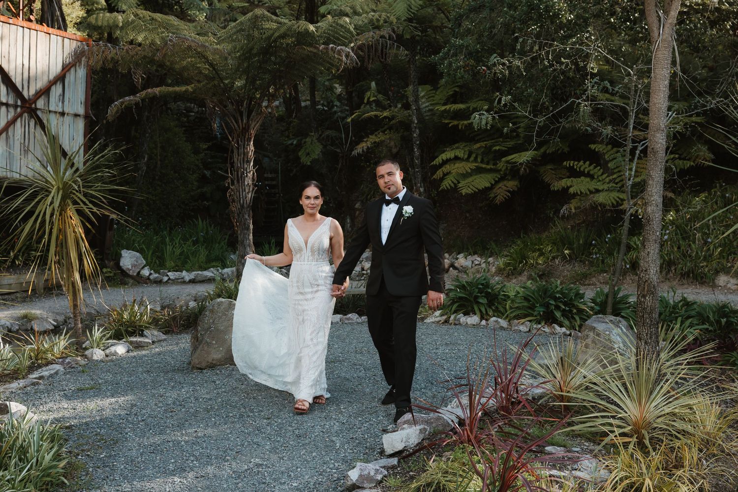 A couple walks together on a gravel garden path surrounded by lush greenery and palm trees on a sunny day.