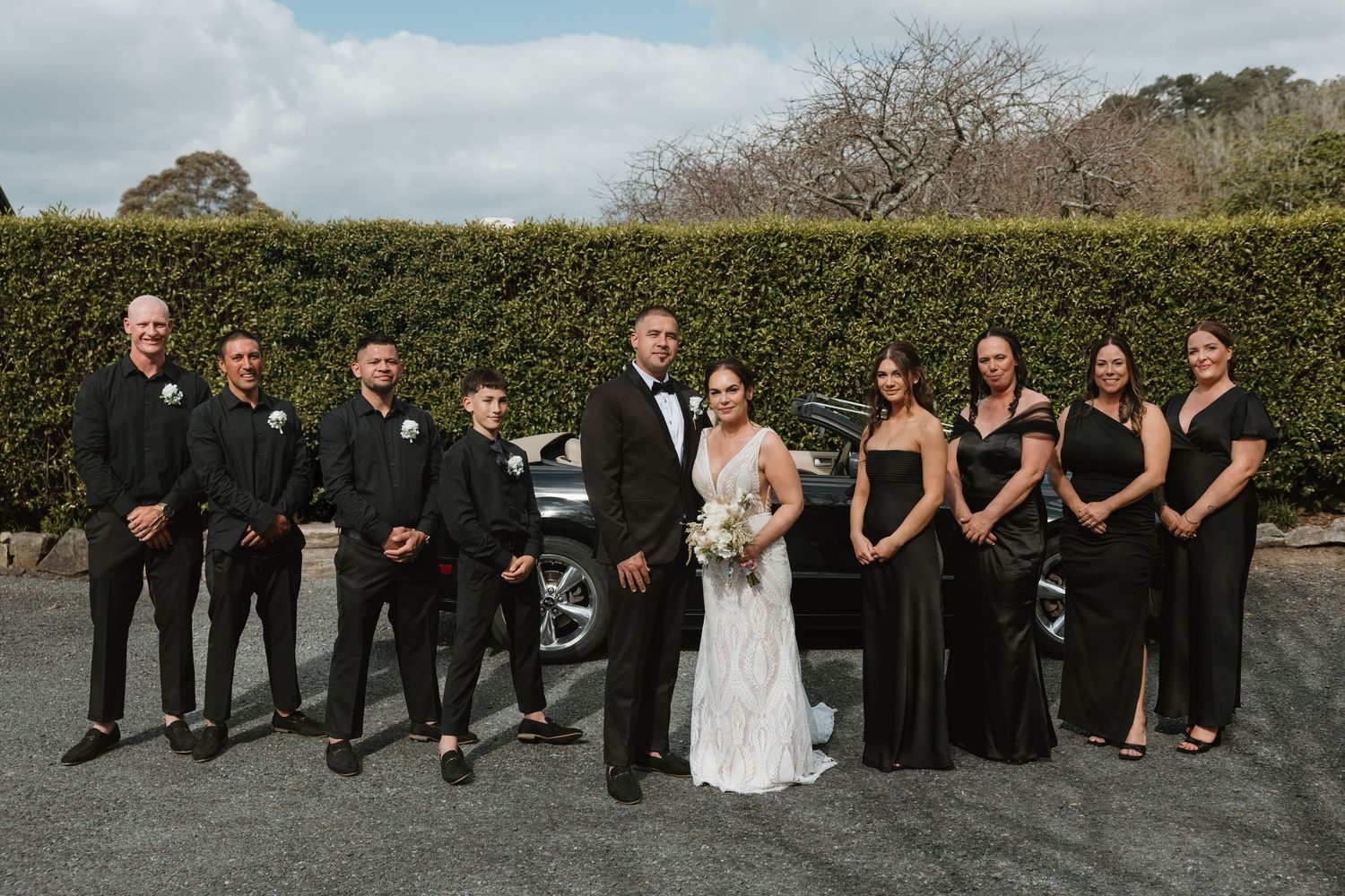 Wedding party in black attire poses playfully on ground while bride in white dress stands smiling.