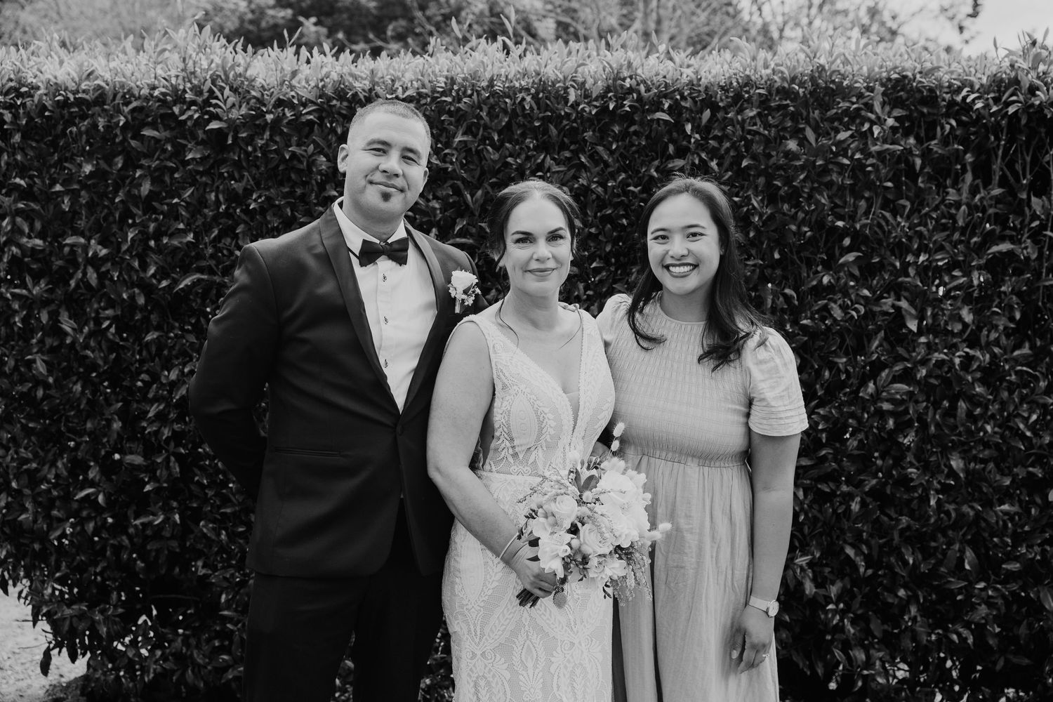 Wedding party members pose together in front of lush green hedges at an outdoor garden venue.