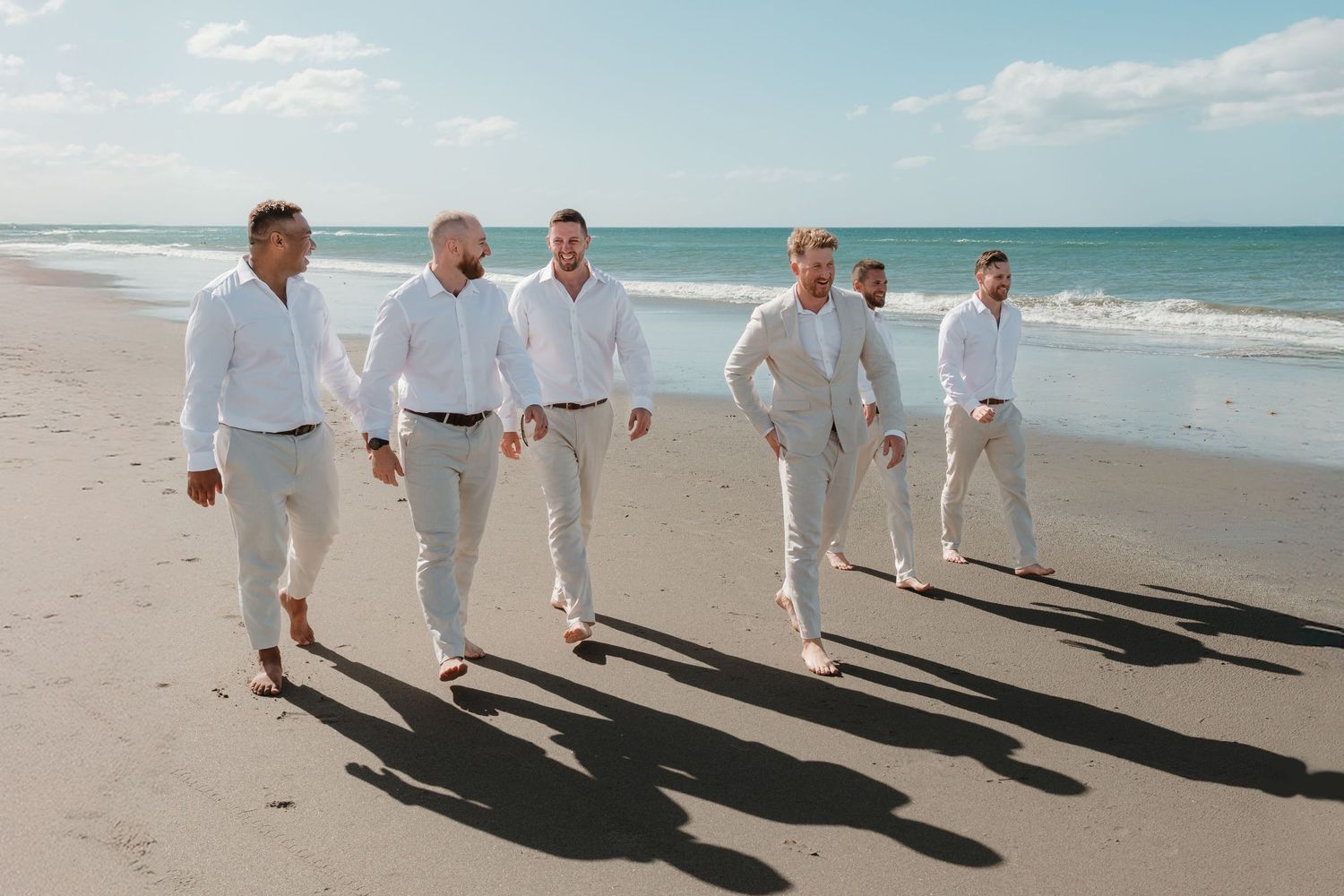 Six groomsmen in white shirts and tan pants walk along a sunny beach, casting long shadows on the sand.