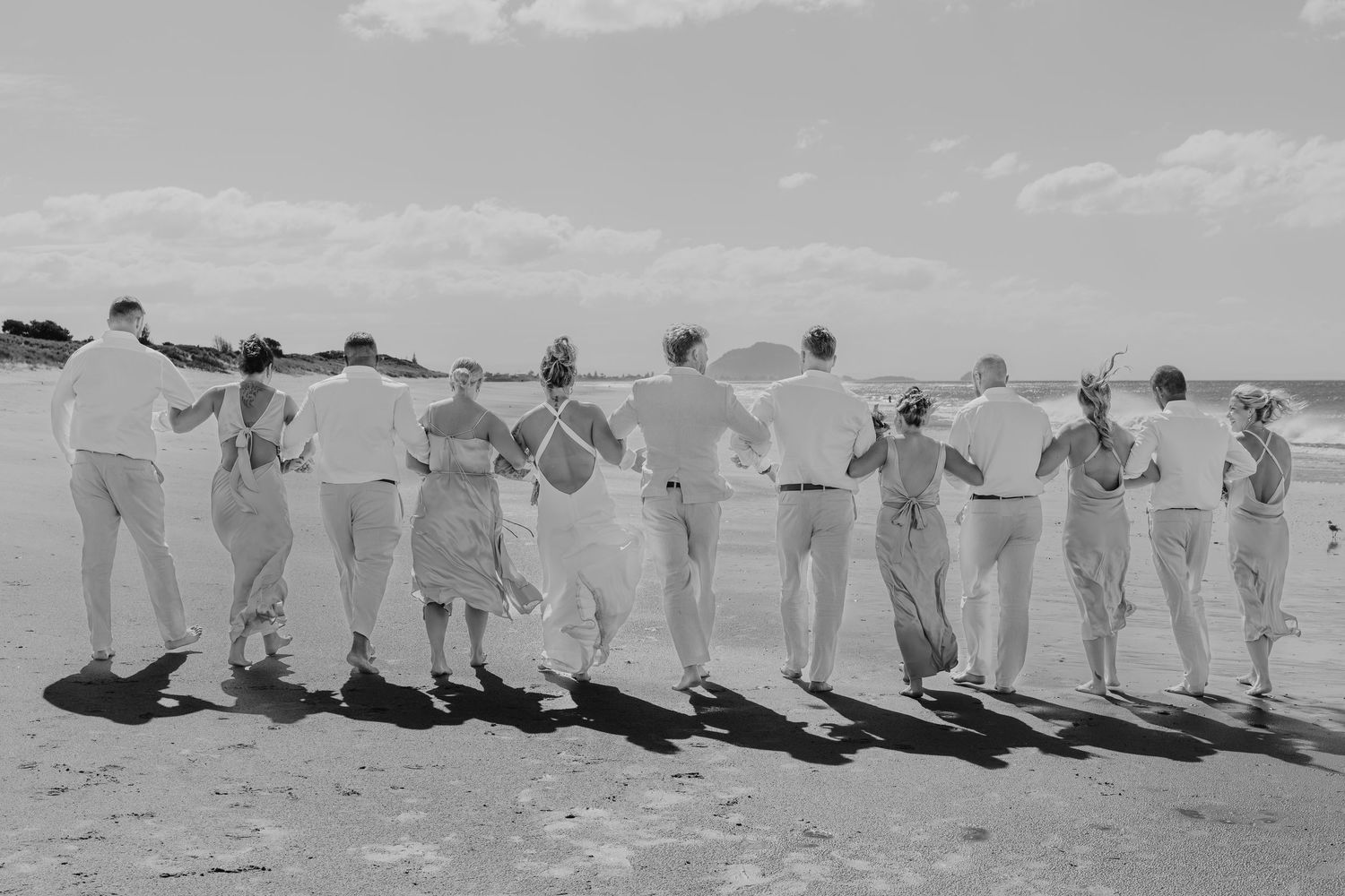 Black and white photo of a wedding party walking arm-in-arm along a sunny beach, backs to camera.
