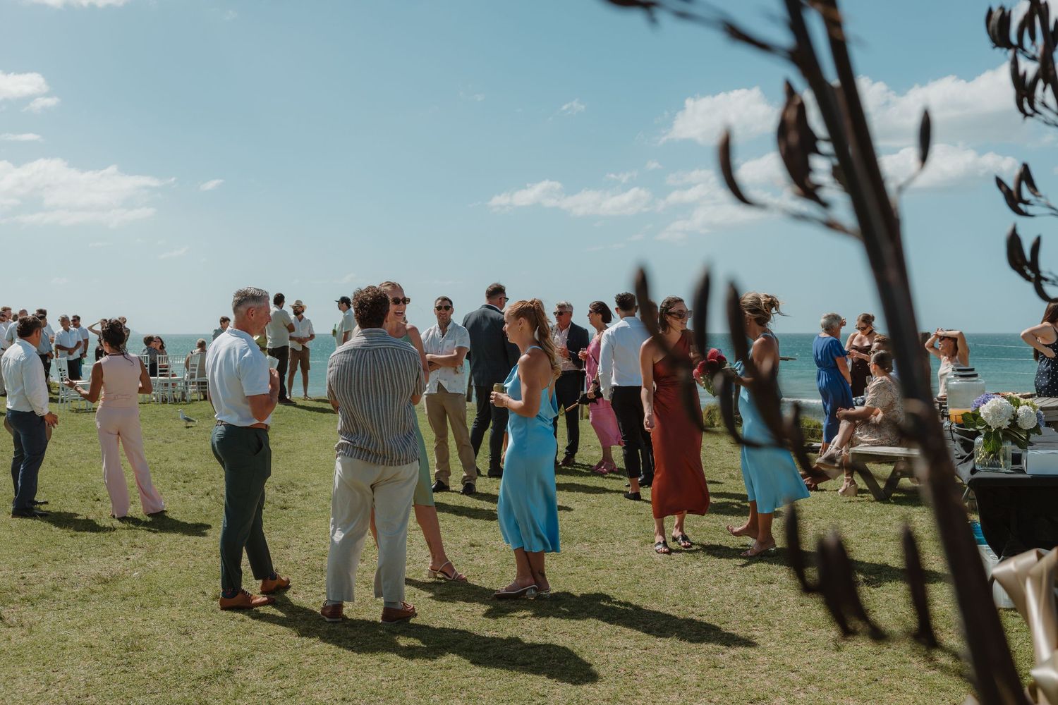 Outdoor coastal wedding reception with guests mingling on green lawn under blue sky near the ocean.