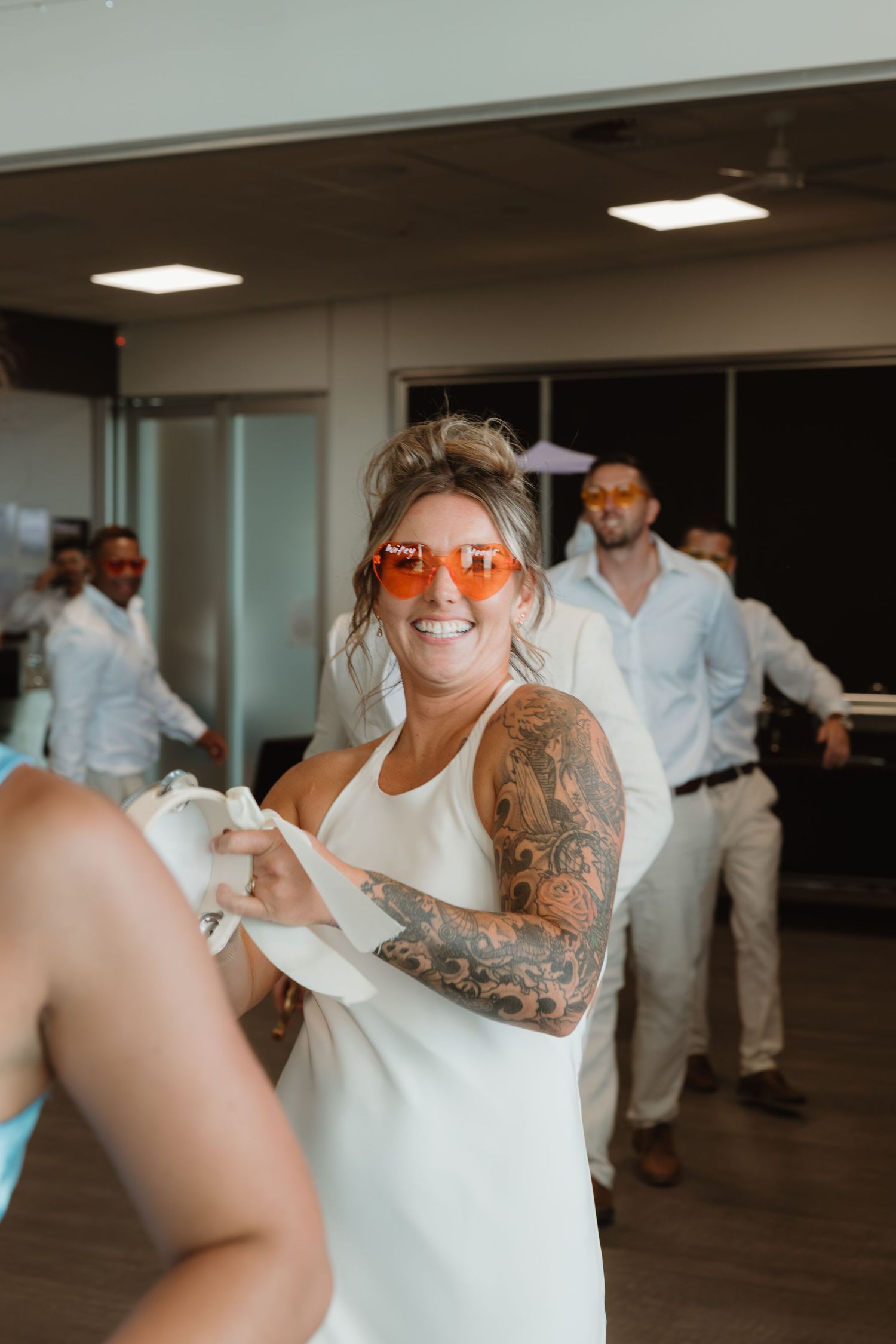Woman in white dress with tattoos and orange sunglasses dancing at a party with guests in background.