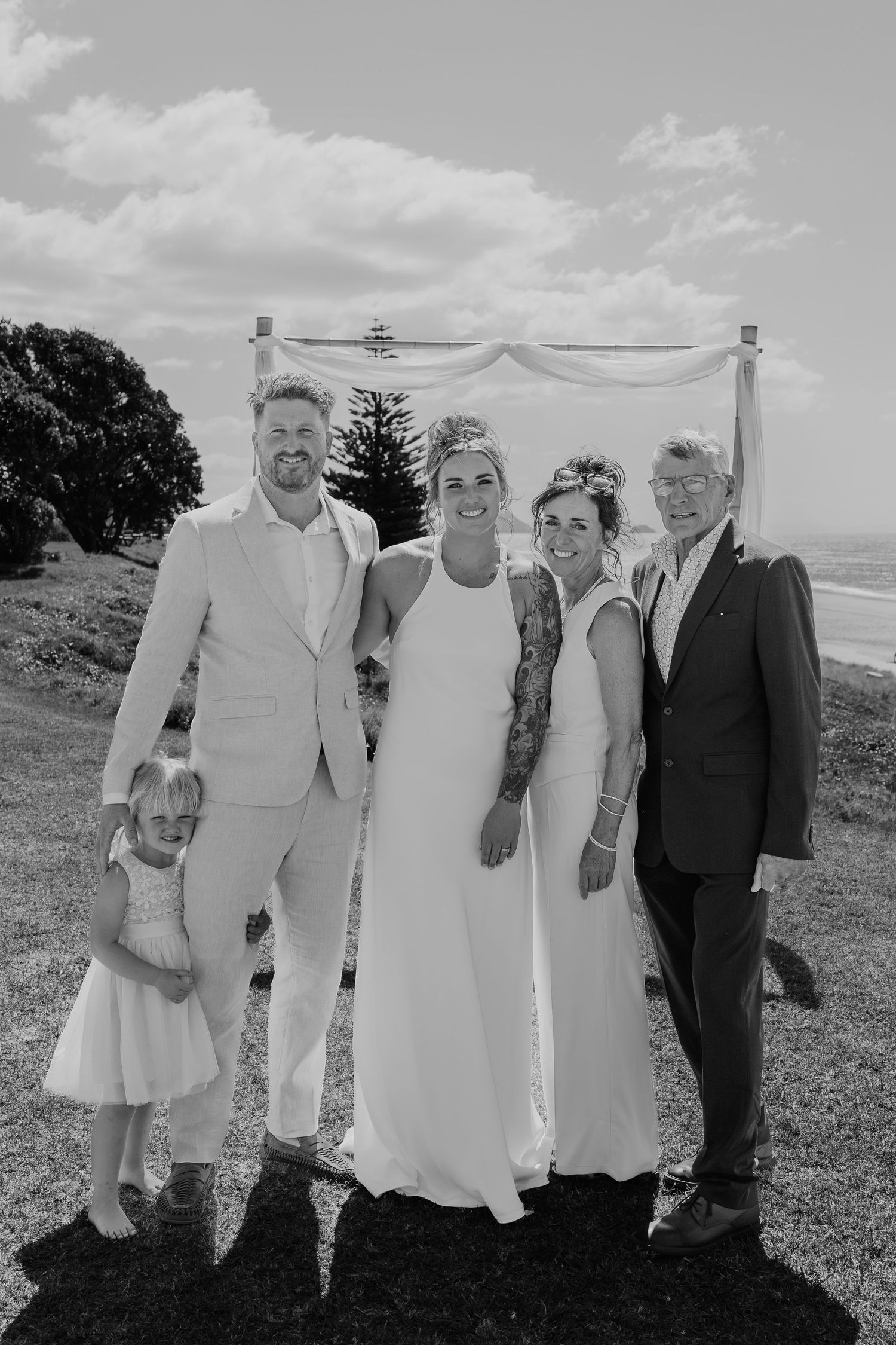 Black and white beach wedding photo of couple with family including young girl in white dress posing before ocean backdrop.