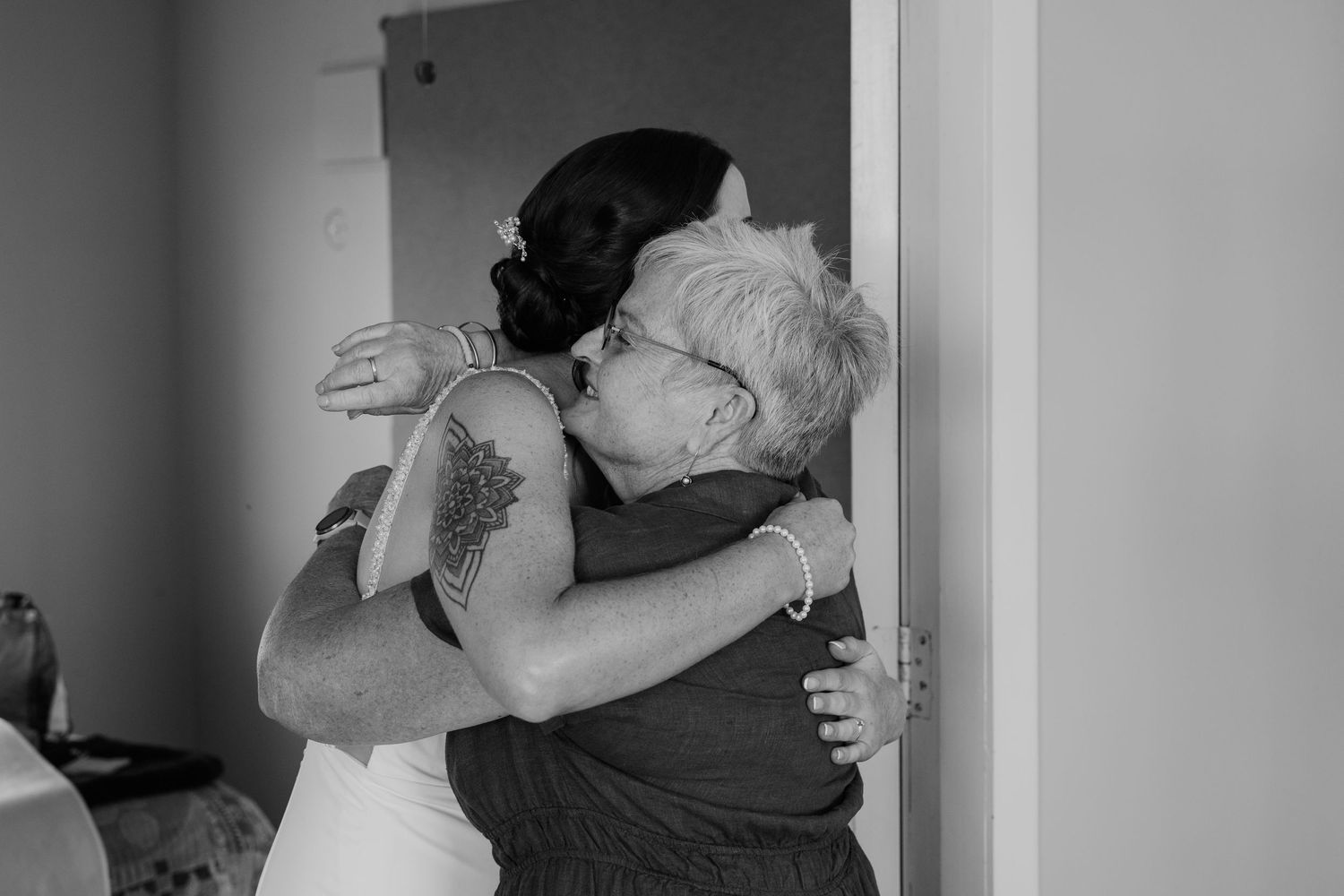 Black and white photo of a bride with tattoo embracing an elderly woman in a heartfelt wedding day hug.
