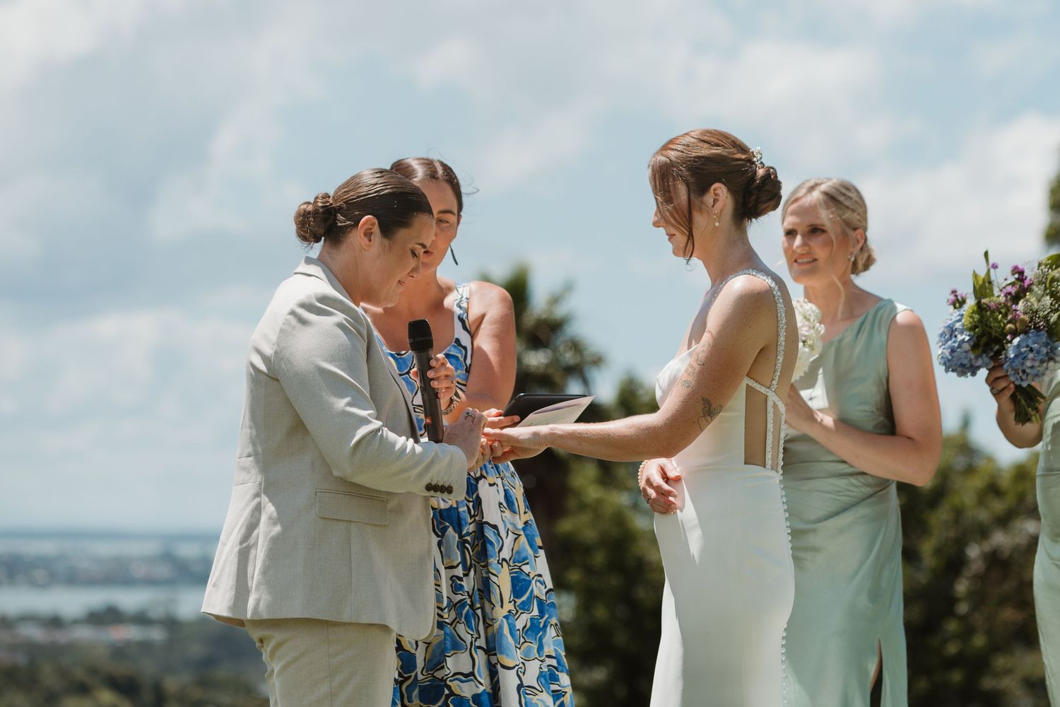 Two brides exchange vows outdoors with an officiant on a sunny day, with scenic views in the background.