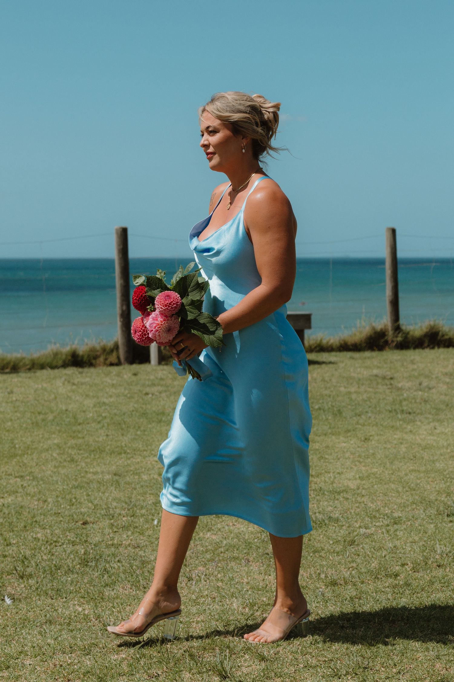 Barefoot bridesmaid in light blue satin midi dress holding pink bouquet walking on grass near ocean.