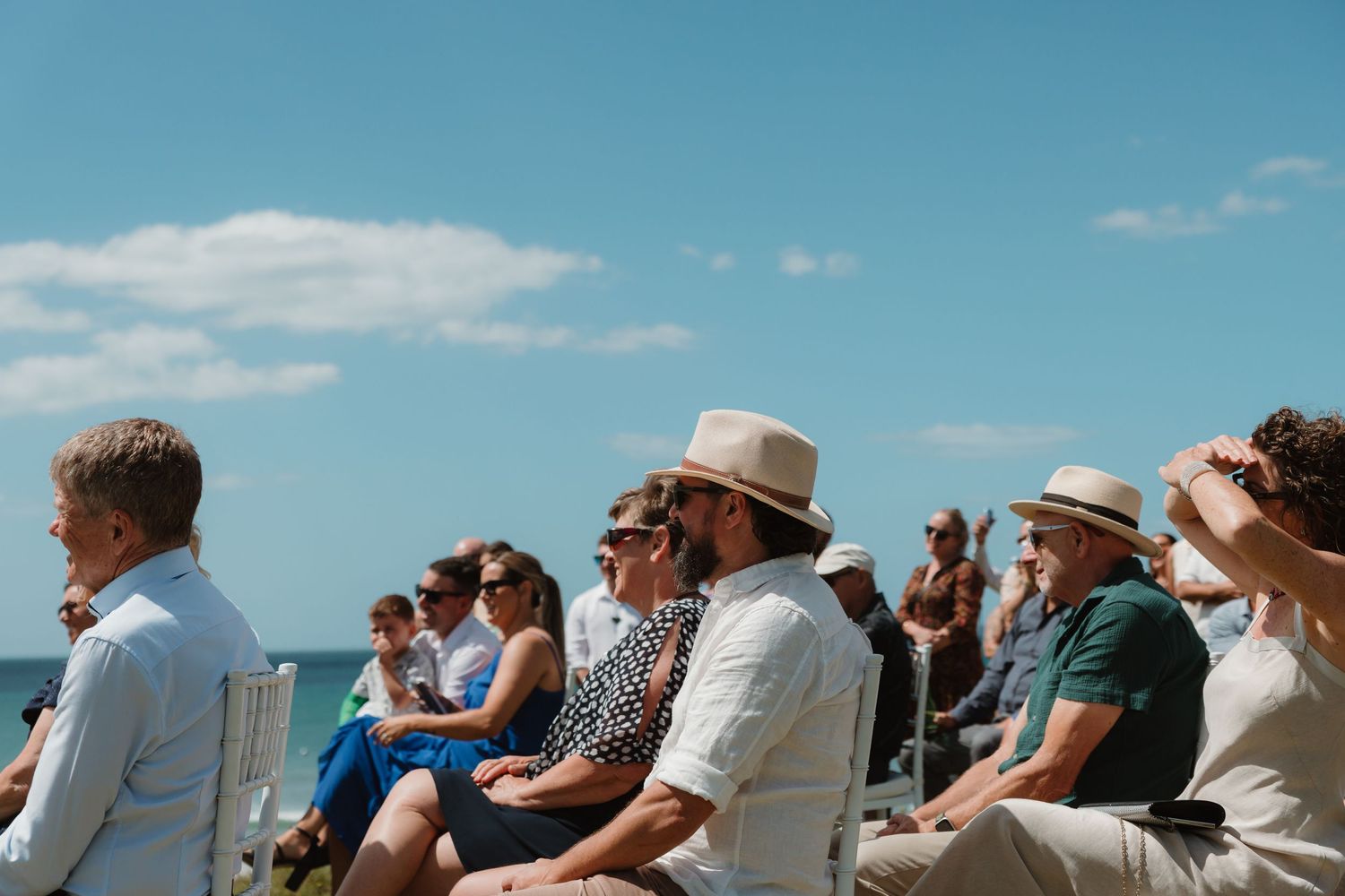 Guests in summer attire and sun hats seated outdoors at a beachside ceremony under a bright blue sky.