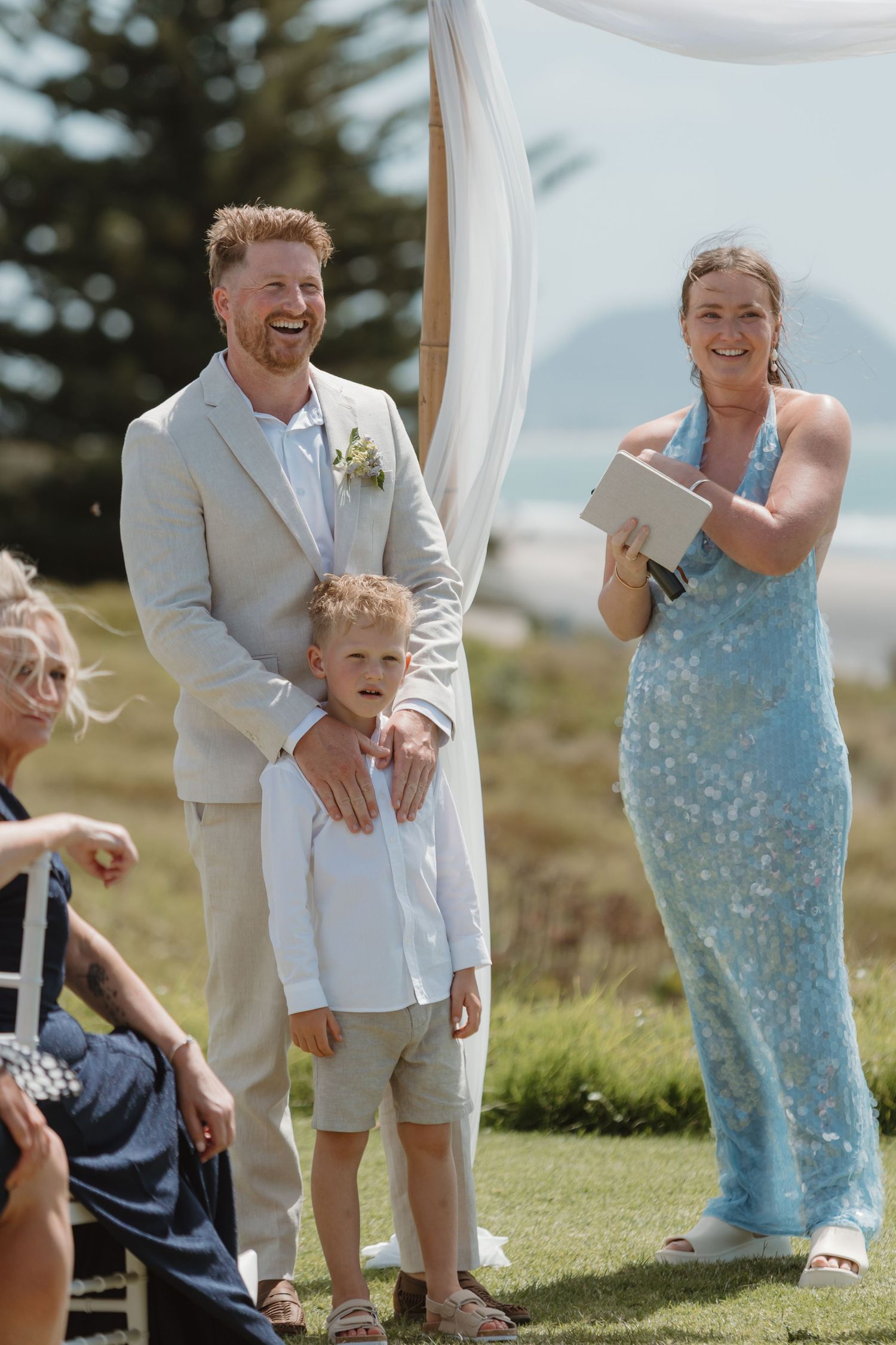 Groom in beige suit laughing with young boy at outdoor beach wedding ceremony, officiant in blue dress holding book.