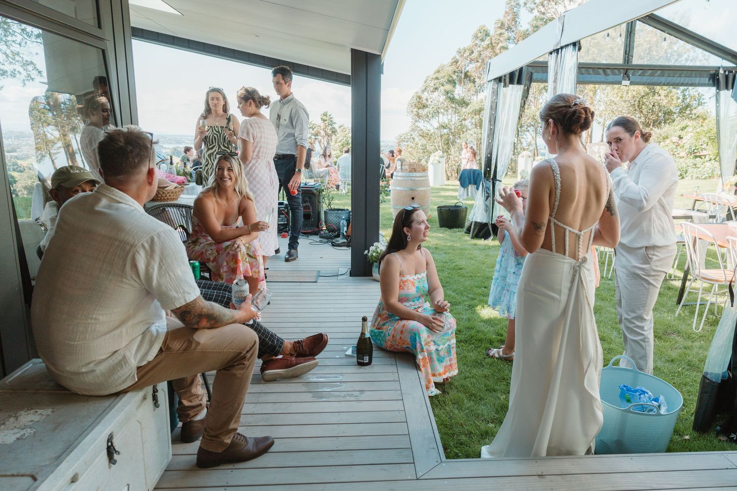 Guests mingling at an outdoor wedding reception on a deck, with a bride in a white gown socializing nearby.