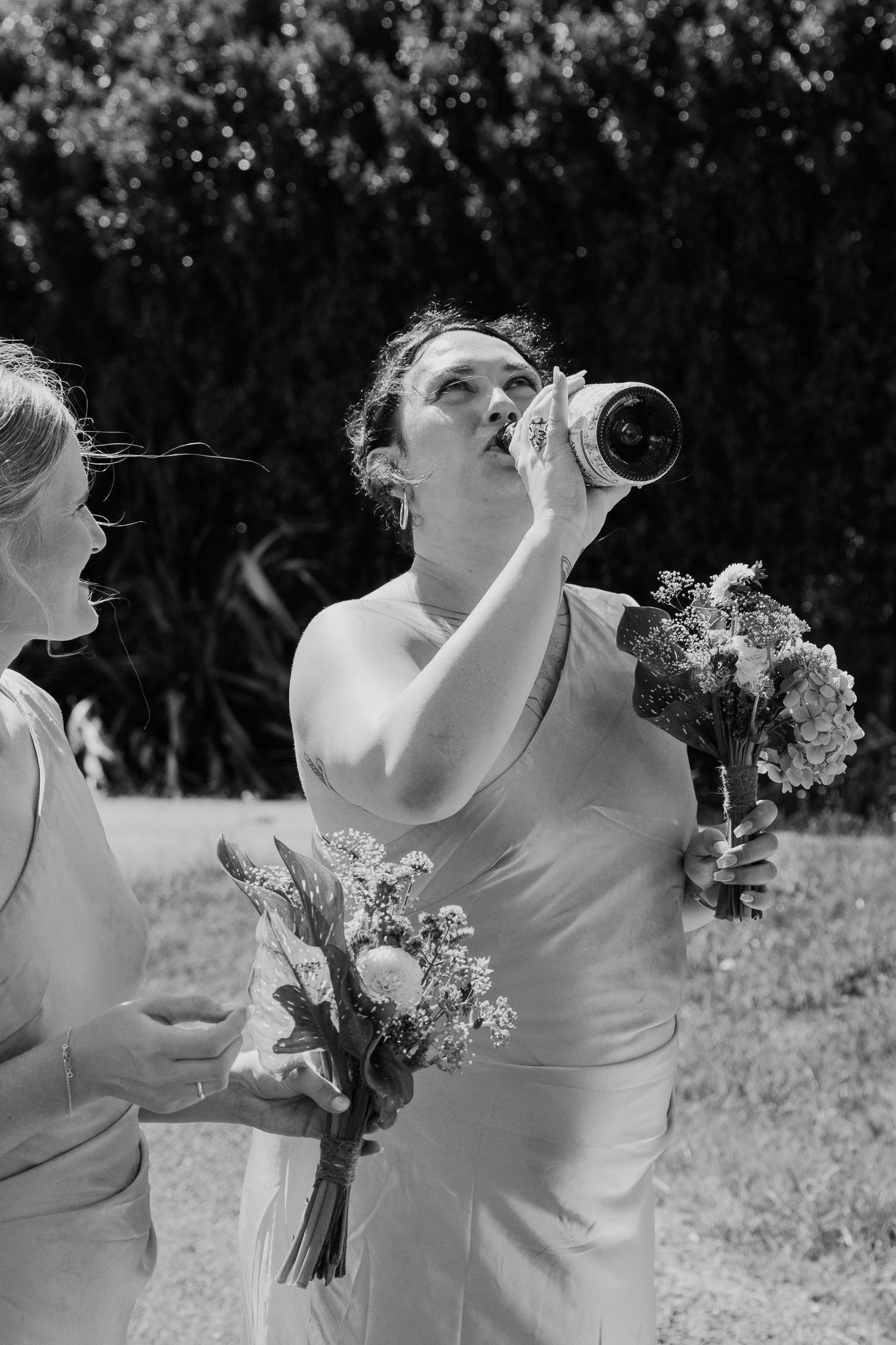 Bride in white gown drinks from wine bottle while holding bouquet, bridesmaid beside her, black and white wedding photo.