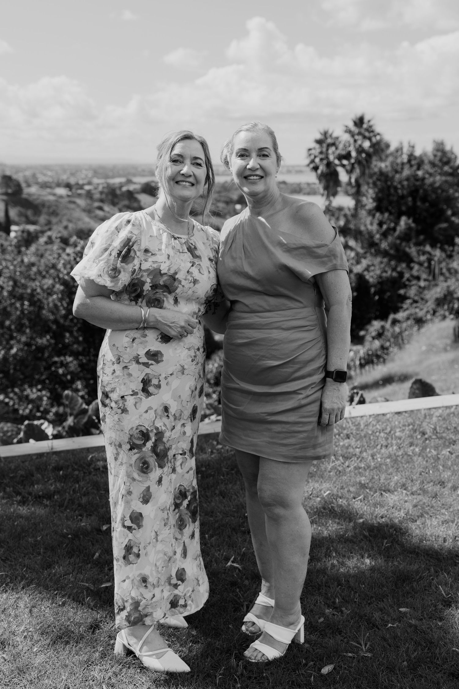 Two women smiling outdoors in elegant dresses at a garden event, black and white photo.