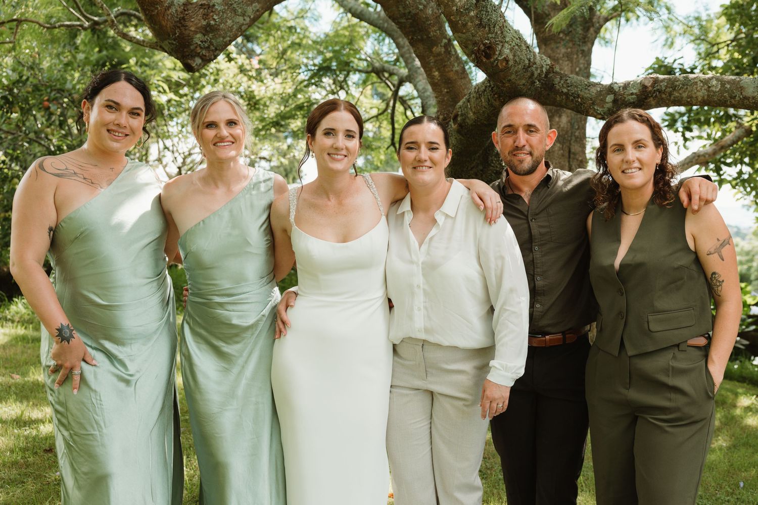 Wedding party posing outdoors under a large tree, featuring a bride in white and guests in sage green attire.