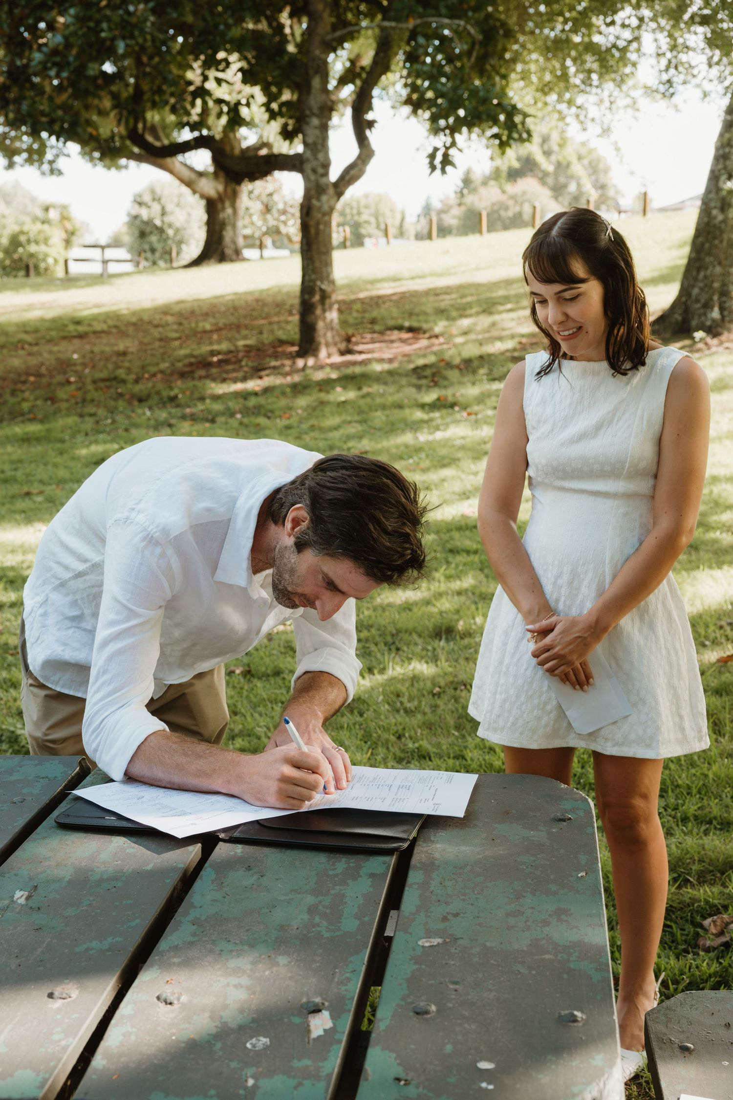 Groom in white shirt signing marriage license at picnic table as smiling bride watches outdoors.