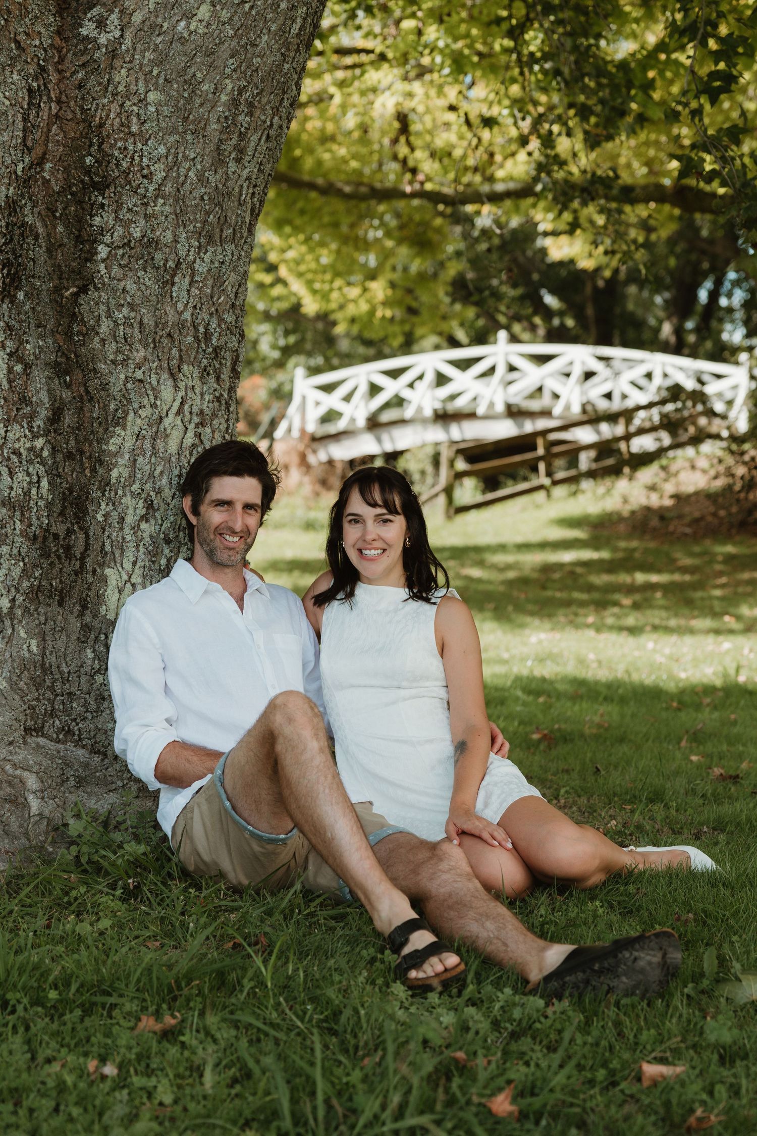 Couple sitting together against a large tree trunk in a lush green park with a white bridge in the background.