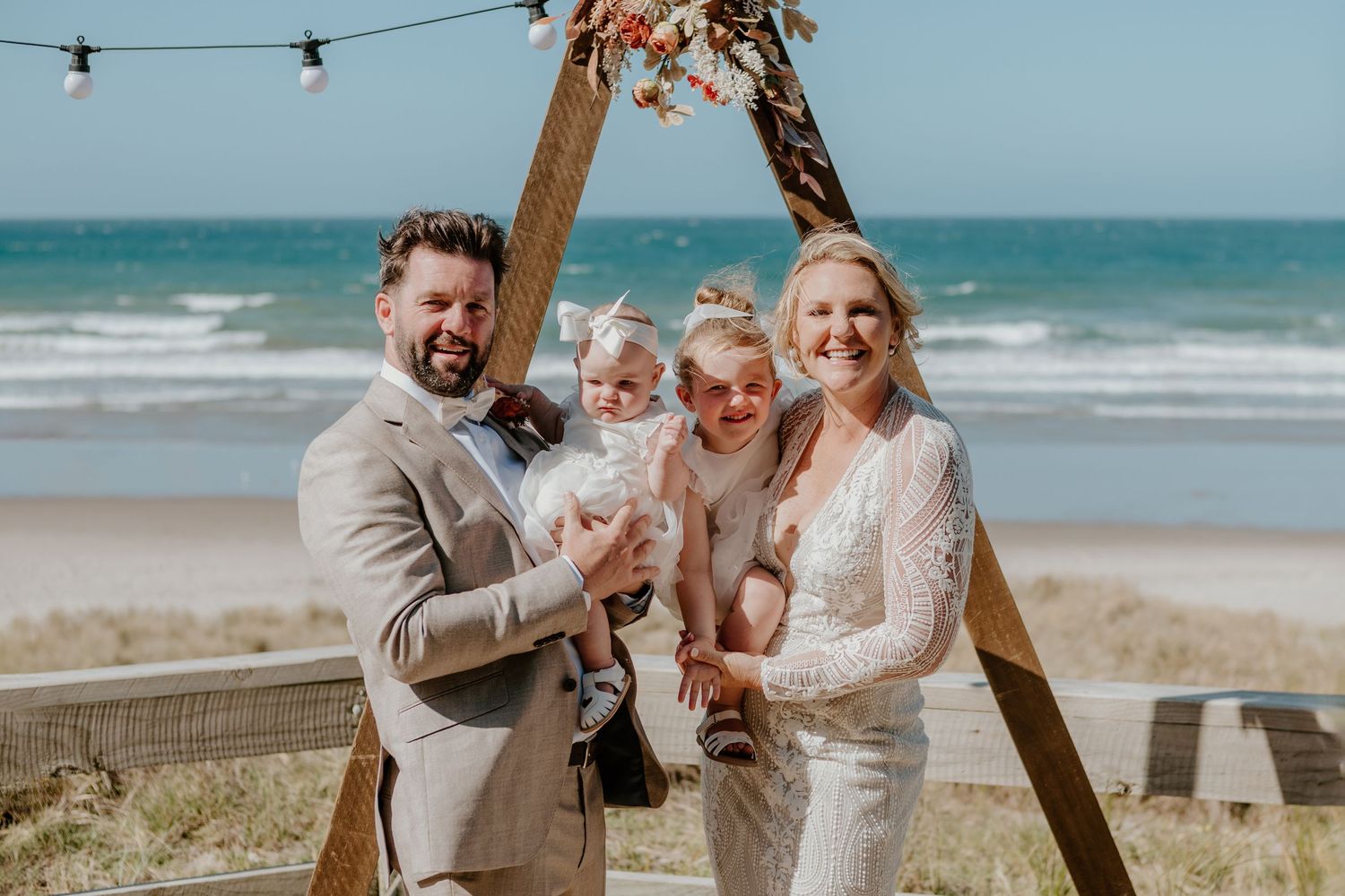 A joyful celebration under a wooden arch at a beach wedding. Papamoa Surf Lifesaving Club Wedding