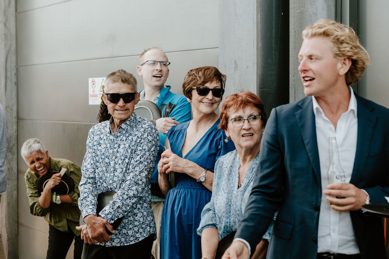 A group of wedding guests smile and laugh while standing together outdoors. Papamoa Surf Lifesaving Club Wedding