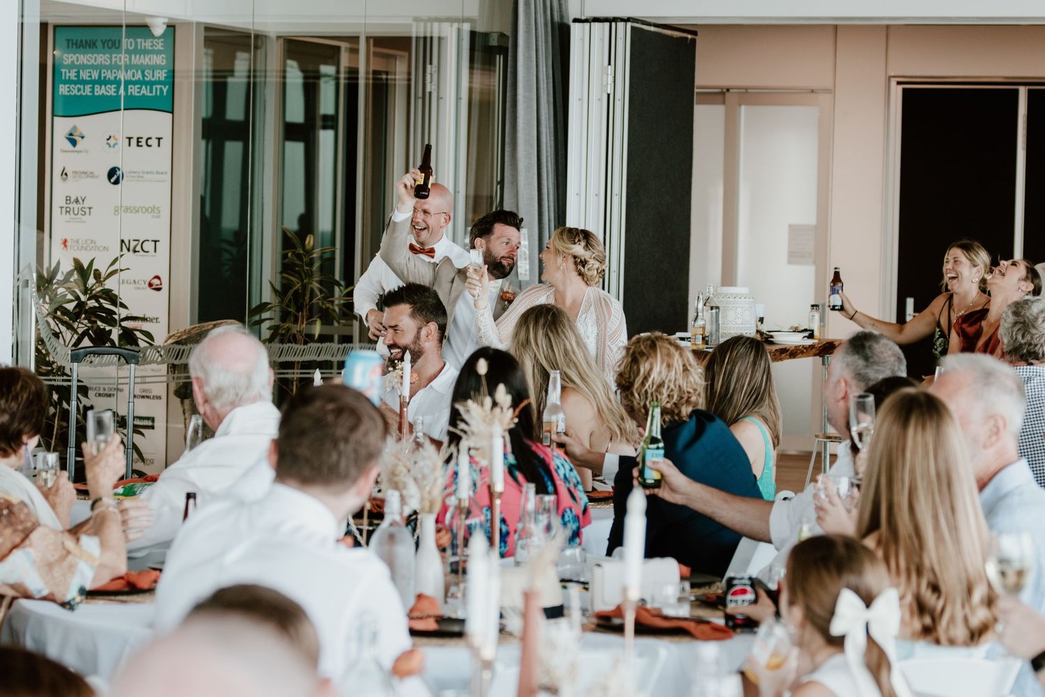 Modern reception venue with white decor and guests seated at long dining tables. Papamoa Surf Lifesaving Club Wedding
