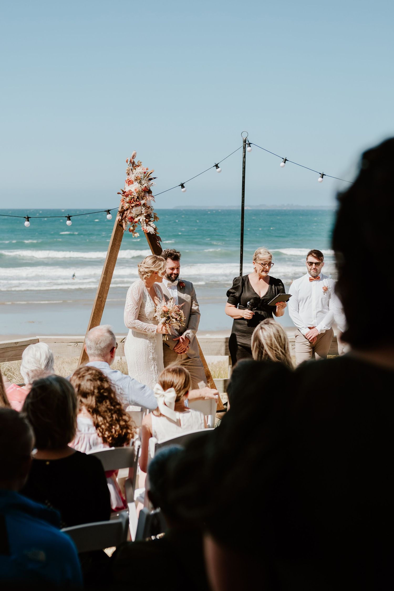 Seaside wedding ceremony with wooden arch and floral decorations against blue ocean backdrop. Papamoa Surf Lifesaving Club Wedding