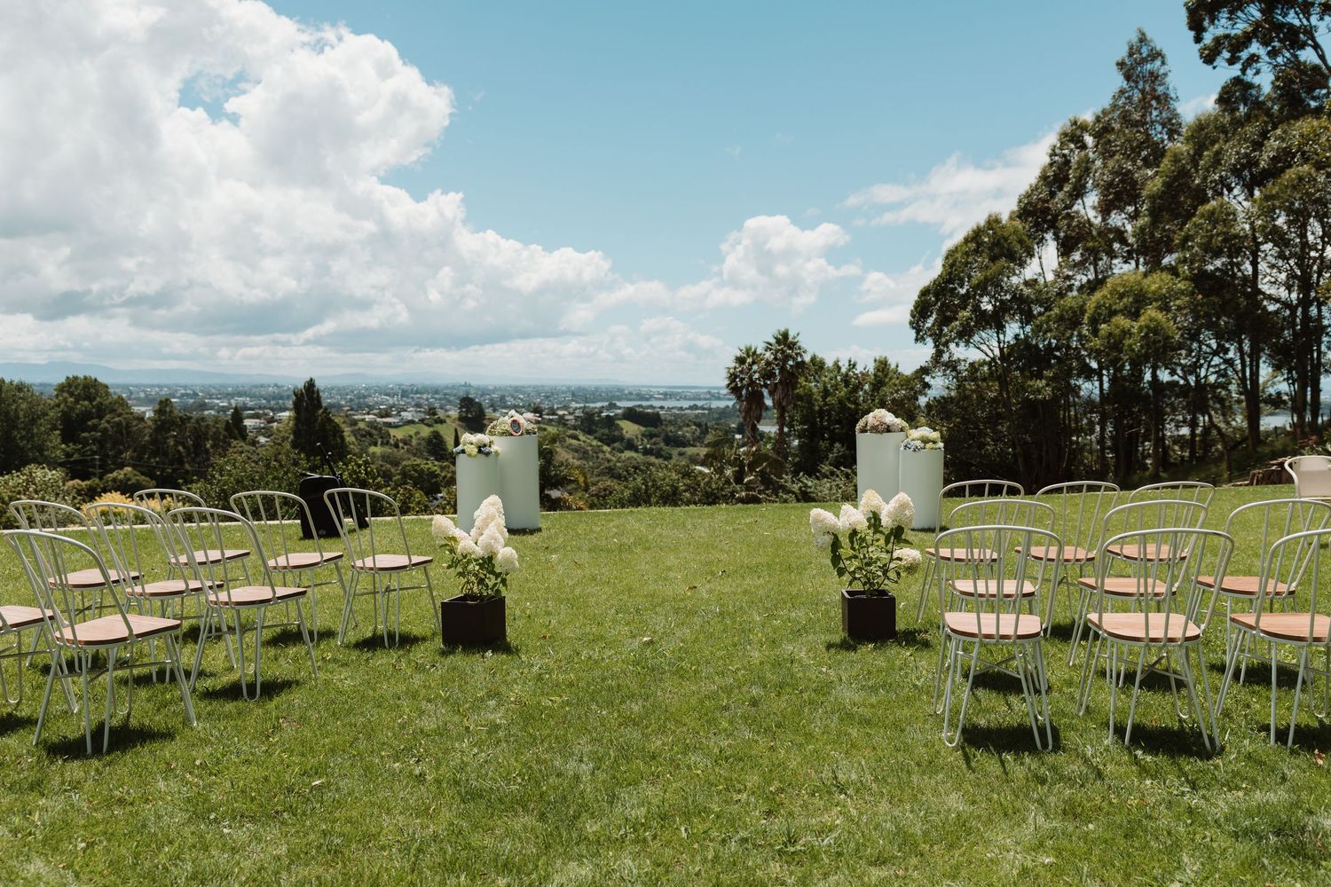 Outdoor wedding ceremony setup on green lawn with white pedestals, wooden chairs, and panoramic countryside views.