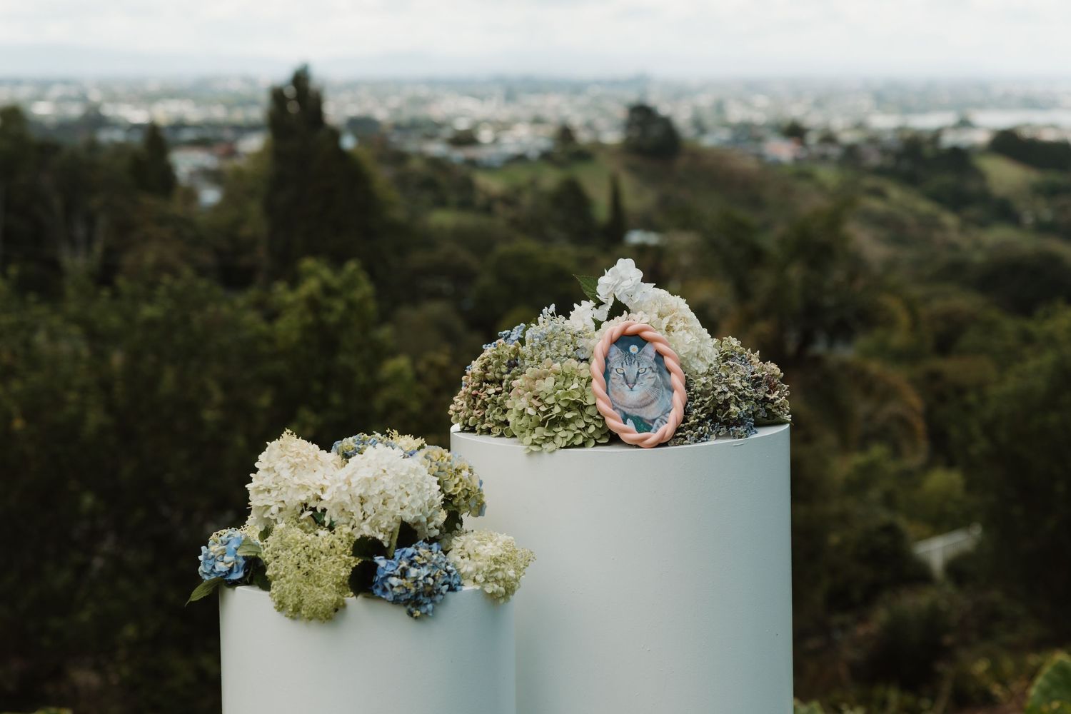 White pedestal with hydrangeas and a geode crystal decoration, set against a lush green landscape and city skyline backdrop.