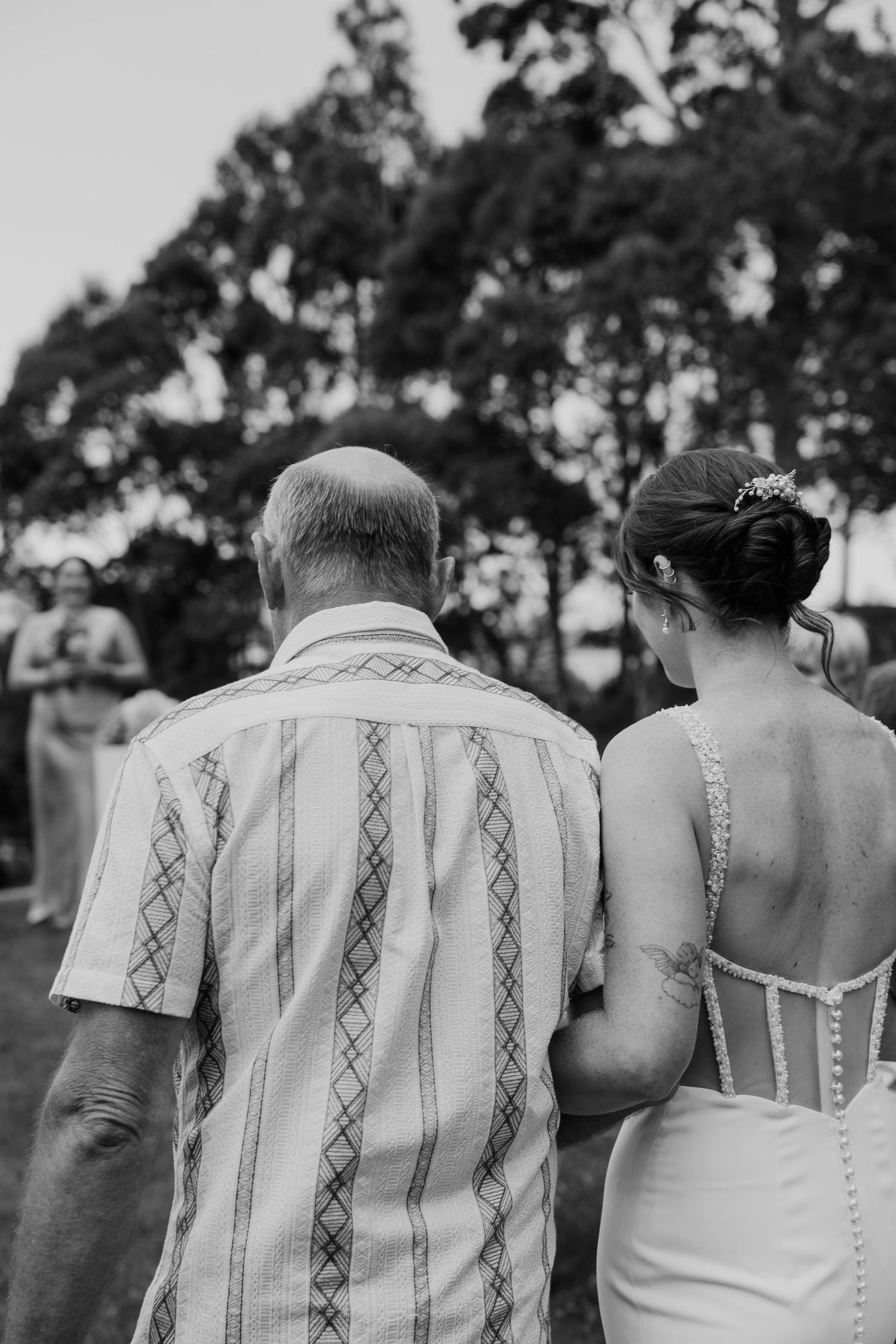 Black and white photo of a bride in white dress walking arm in arm with an older man at an outdoor wedding.