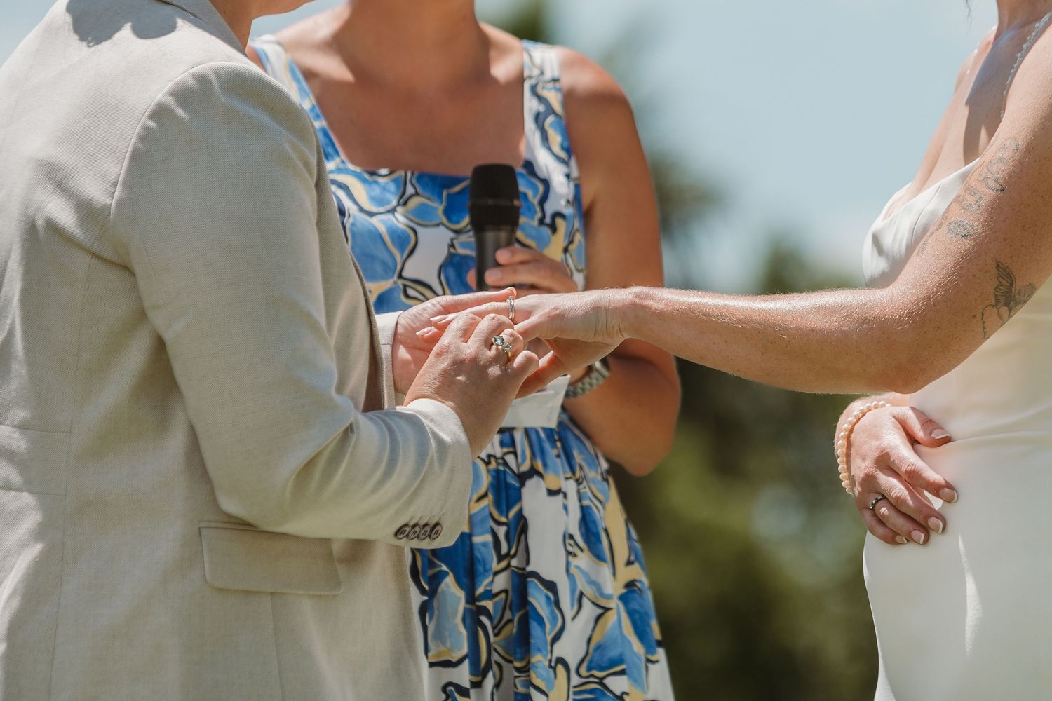 Couple exchanging rings during an outdoor wedding ceremony with officiant in blue floral dress.