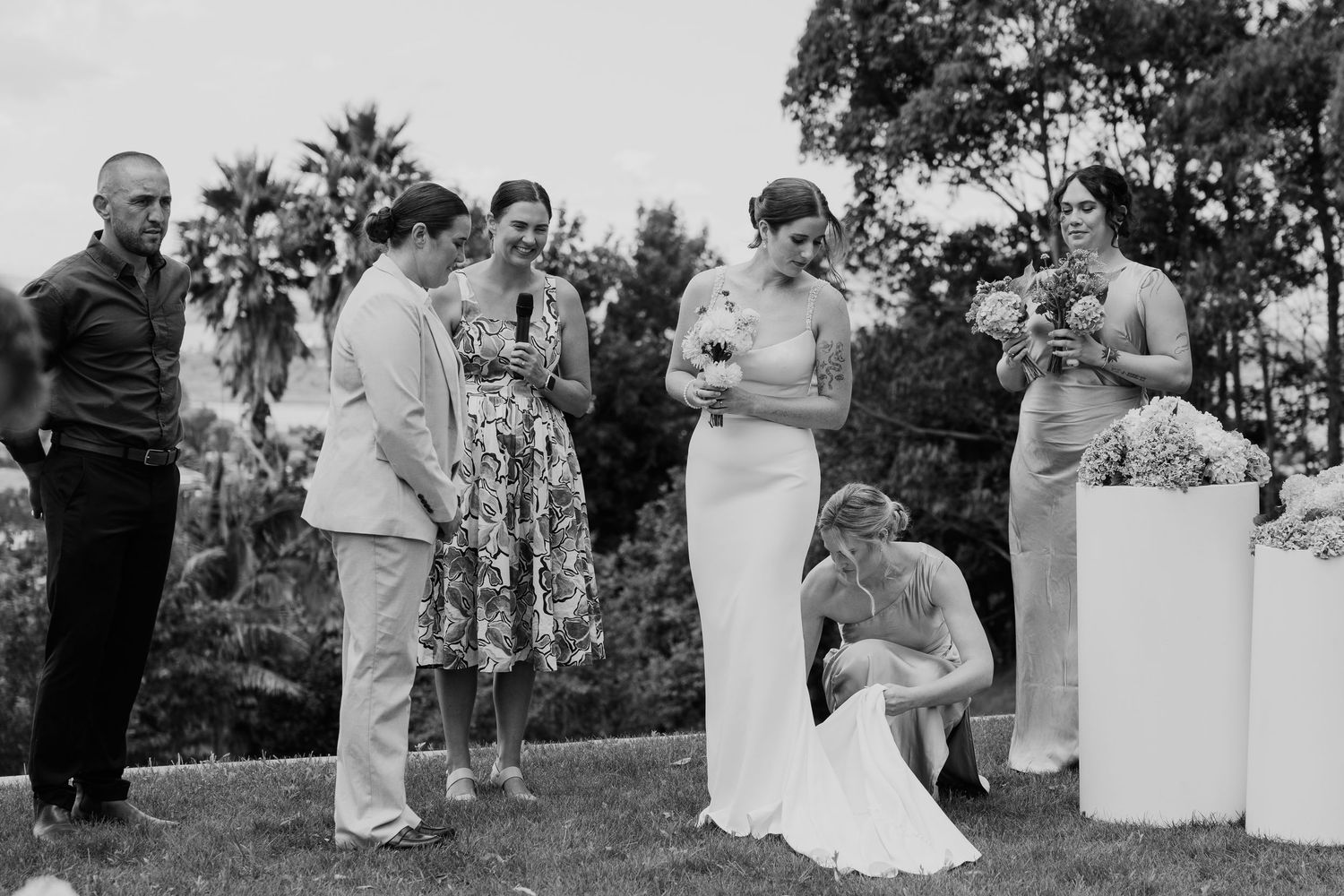 Black and white outdoor wedding ceremony with bride in white gown, bridesmaids, and guests on a lush green lawn.