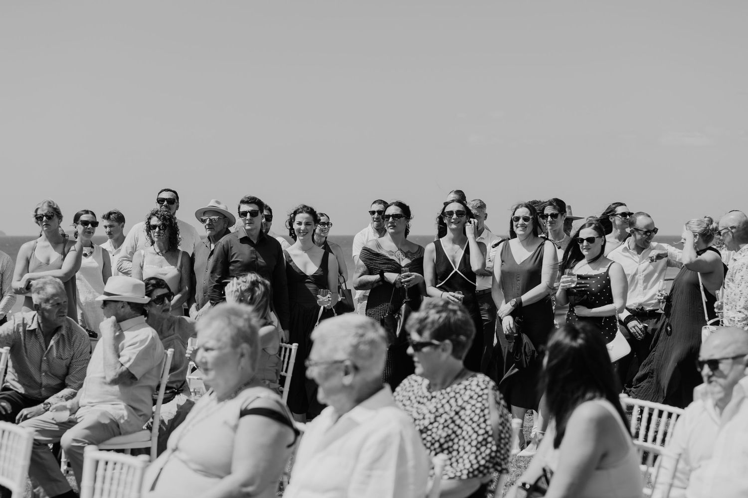 Black and white photo of wedding guests gathered outdoors under clear sky, some seated, others standing in elegant attire.
