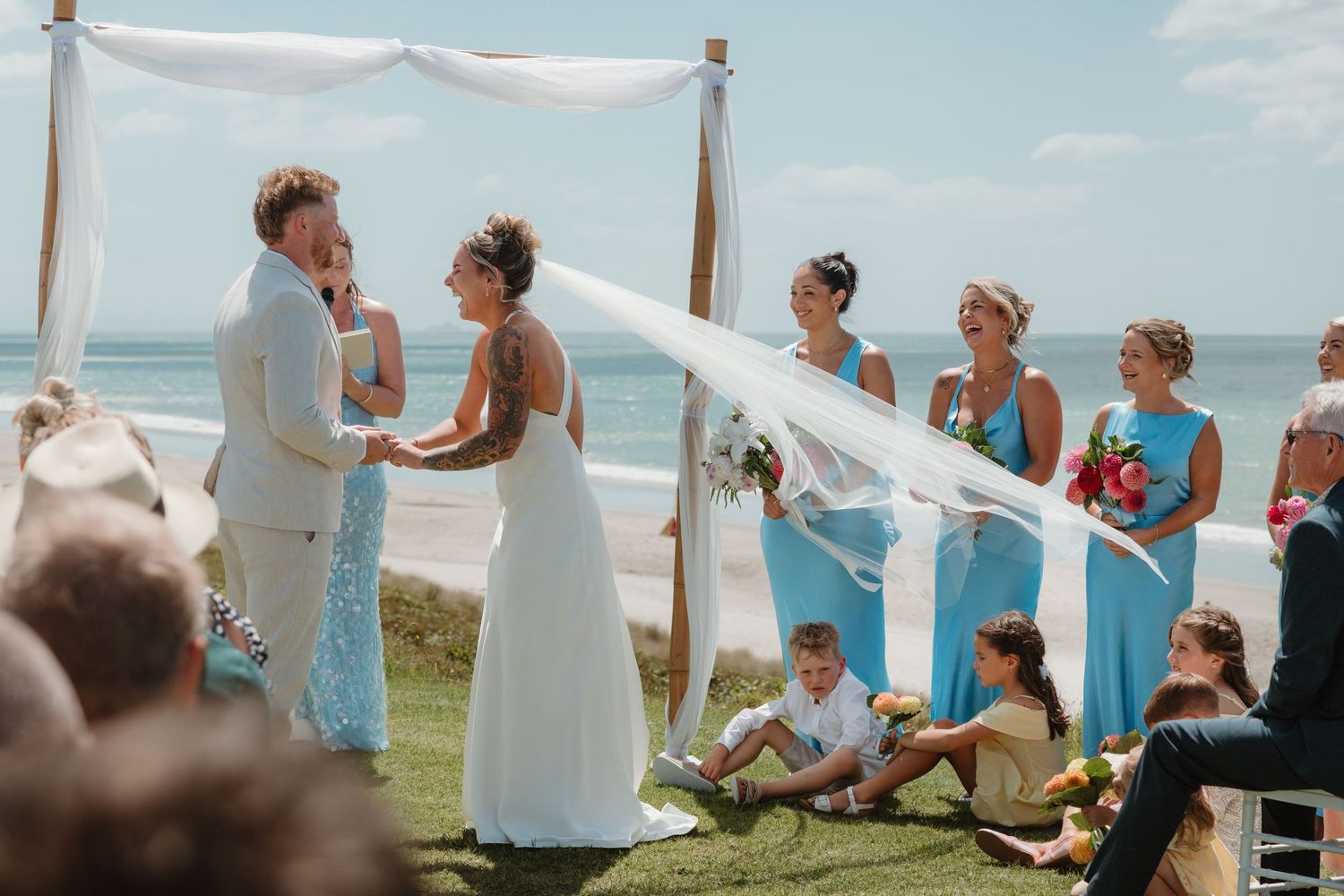 Bride and groom exchange vows at outdoor beach wedding with bridesmaids in blue dresses and ocean backdrop.